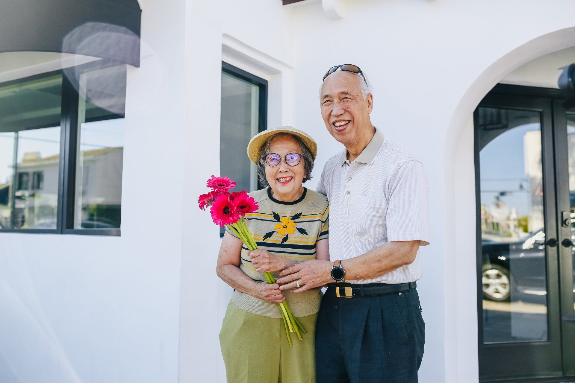 An elderly couple is standing in front of a white building holding flowers.