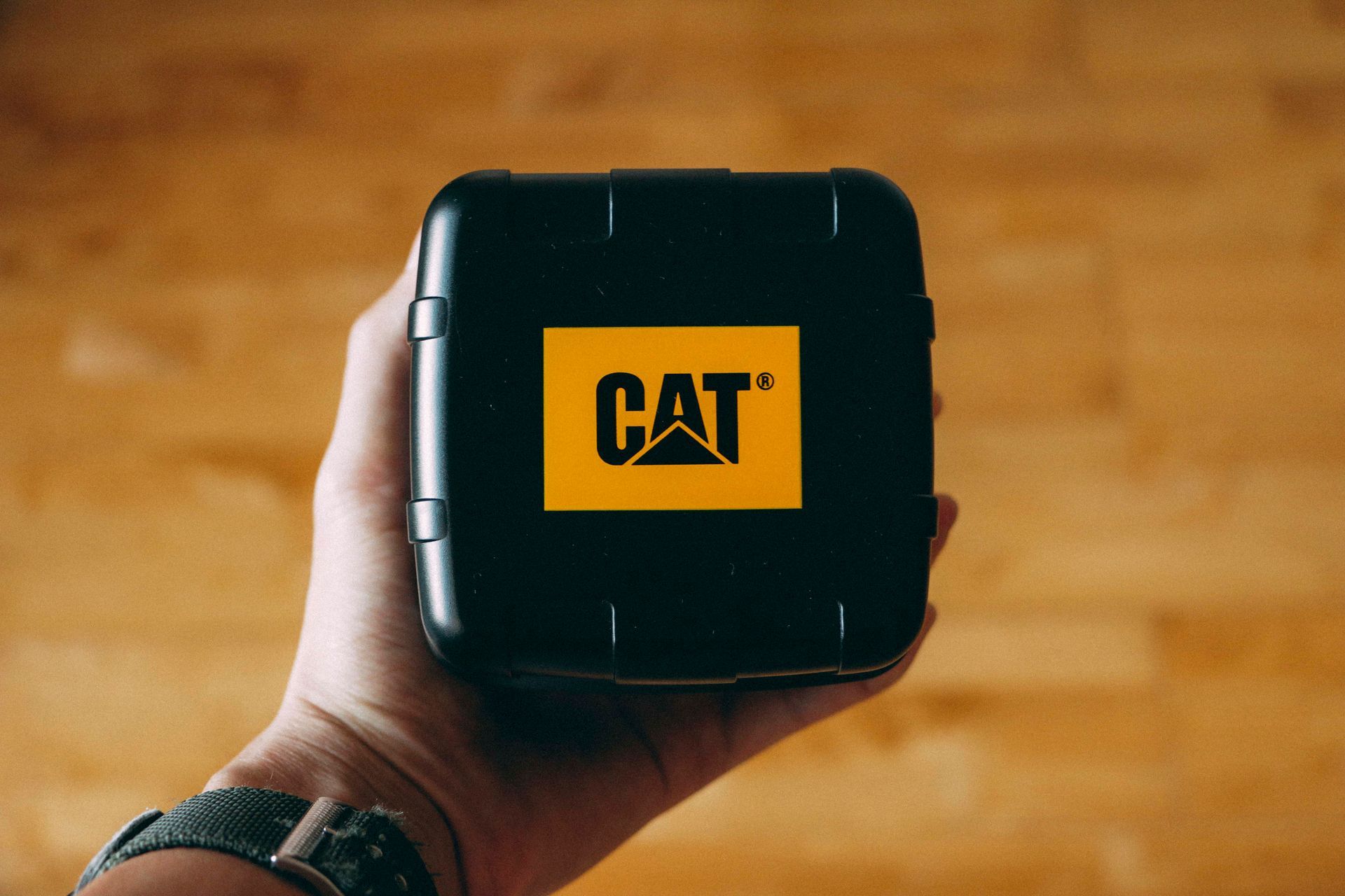 Hand die een zwarte koffer met CAT-logo vasthoudt boven een houten vloer.