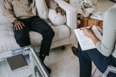 A man is sitting on a couch talking to a woman who is holding a clipboard.