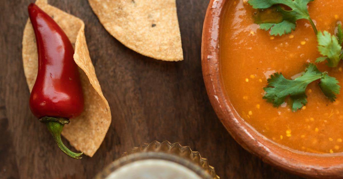 A bowl of soup , tortilla chips , a red pepper and a glass of beer on a wooden table.