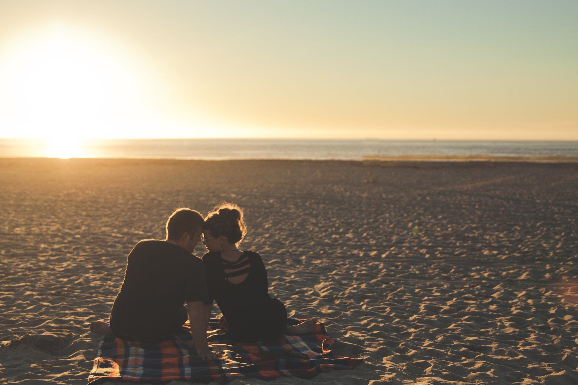 a couple in their thirties sits on a blanket on a beach to watch a sunset together