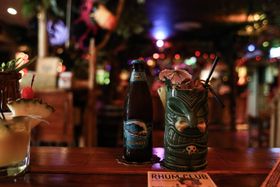 Bar counter with tiki-style cocktails and beer bottle, blurry background of bar interior with lights.