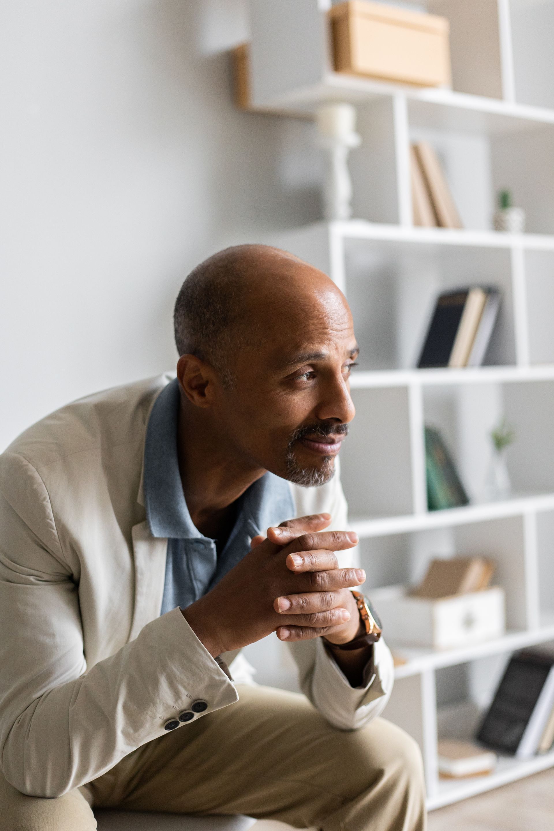 A man is sitting on a stool with his hands folded in front of a bookshelf.