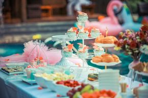 Dessert table with cupcakes, fruit, and pastries beside a pool, decorated in pink and teal.