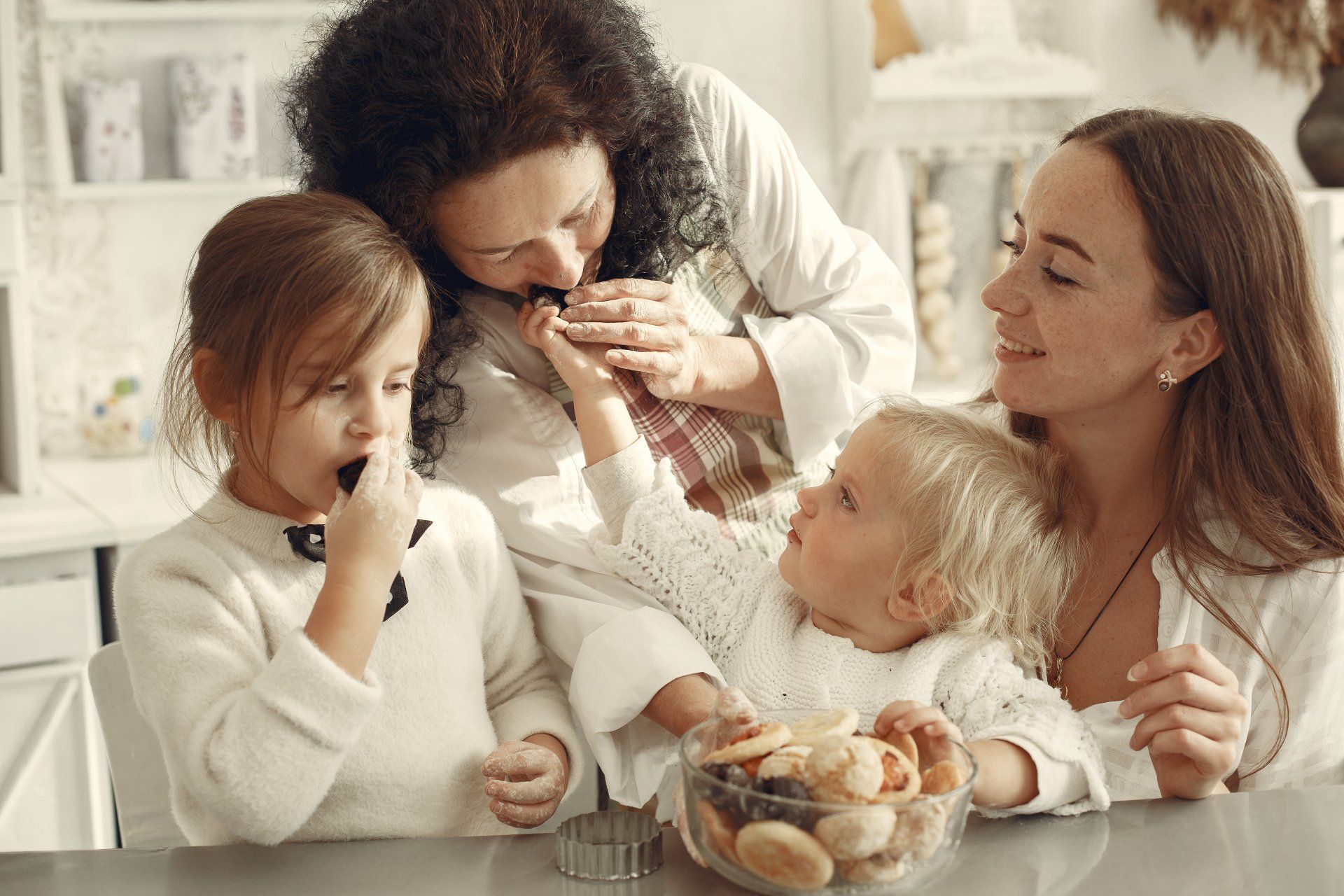 Three women and two children are sitting at a table eating cookies.