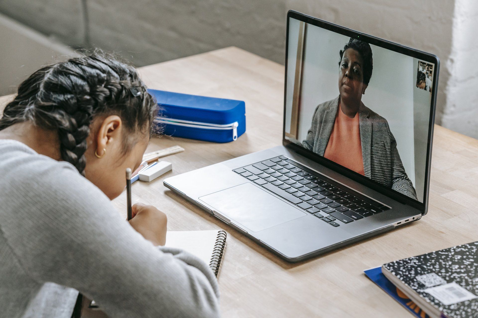 Girl with braids, studying with a laptop open showing a video call with a teacher.