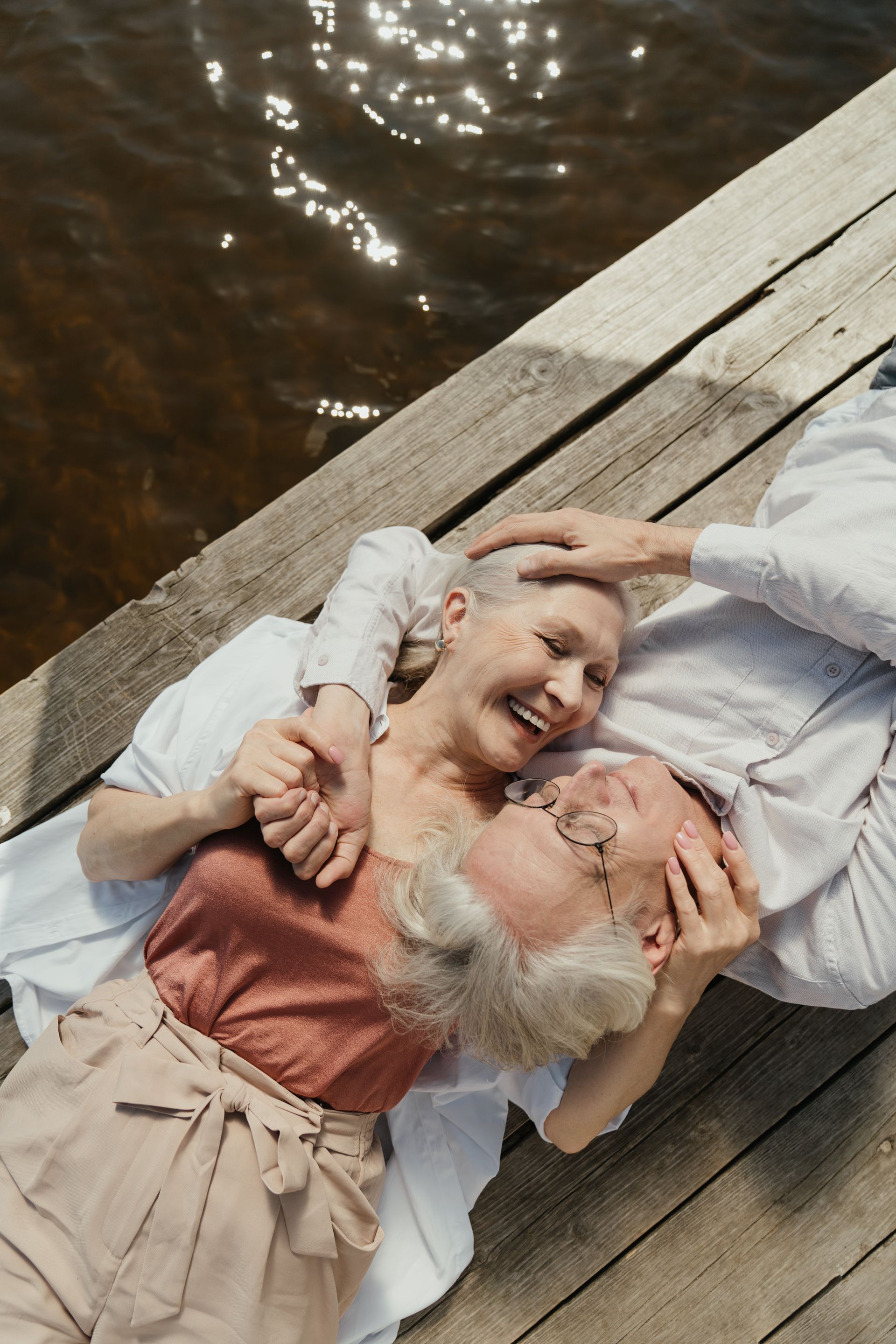 An elderly couple is laying on a wooden dock near the water.