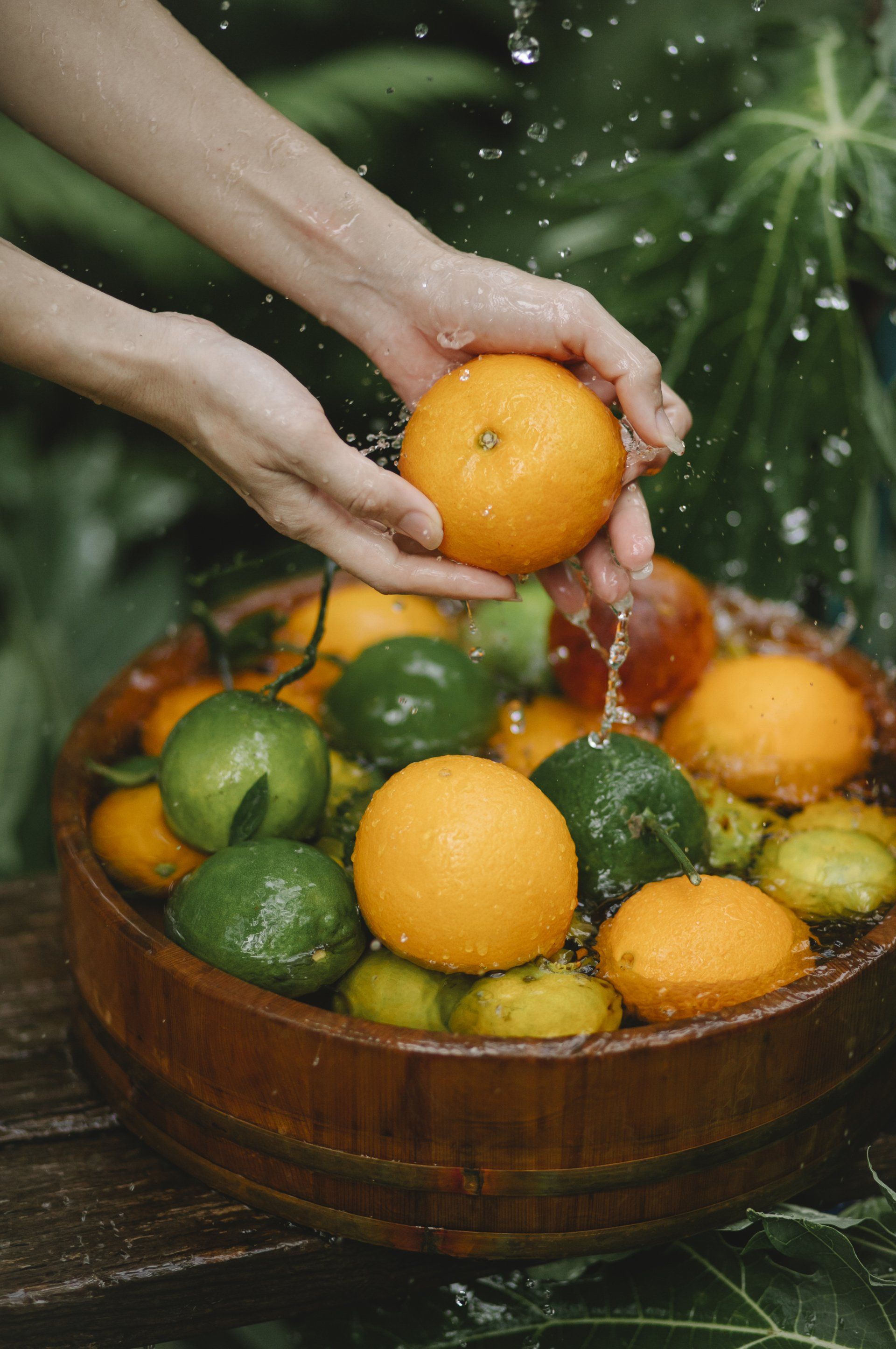 A person is washing oranges and limes in a wooden bowl.