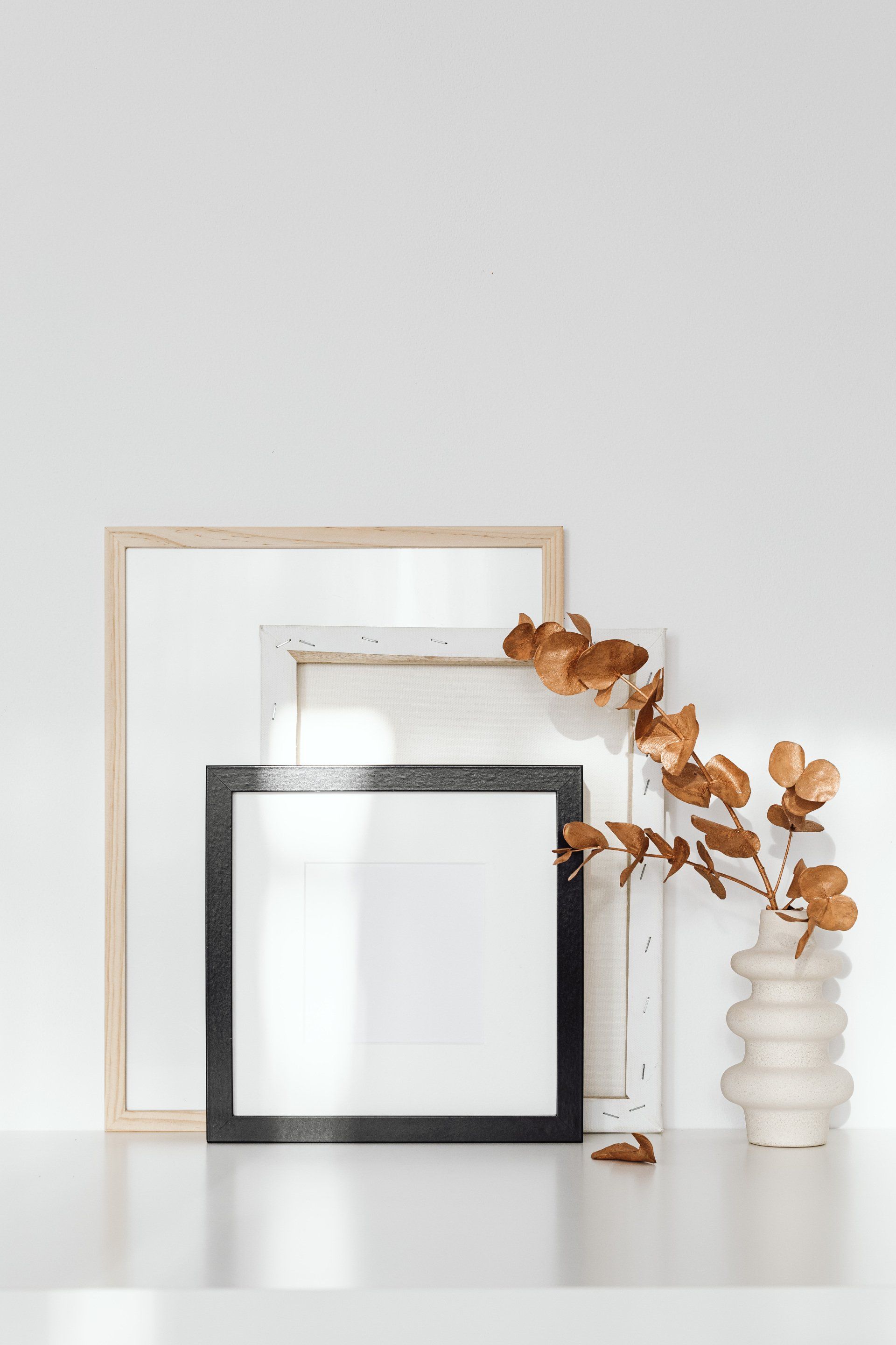 Two picture frames, one black, one white, behind a larger light wood frame. A vase with dried leaves.