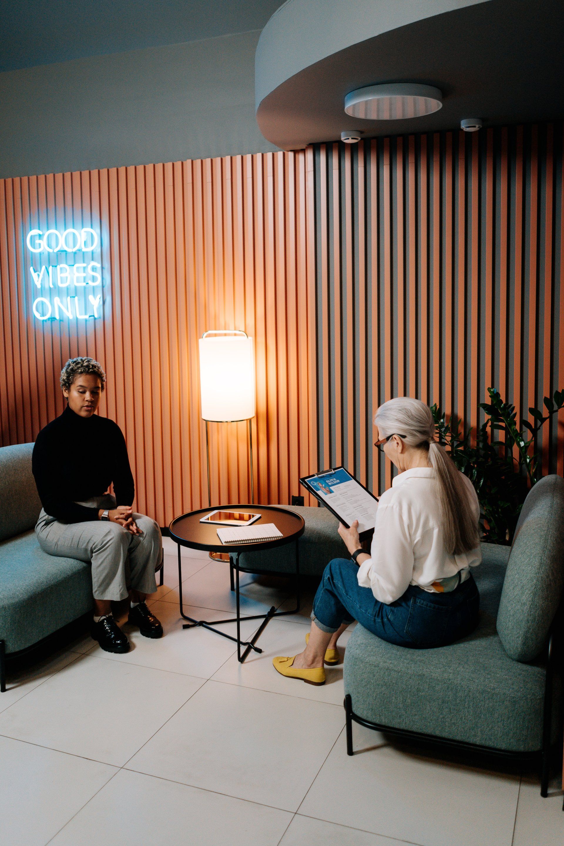 Two women are sitting at a table in a room with a neon sign that says `` good vibes only ''.