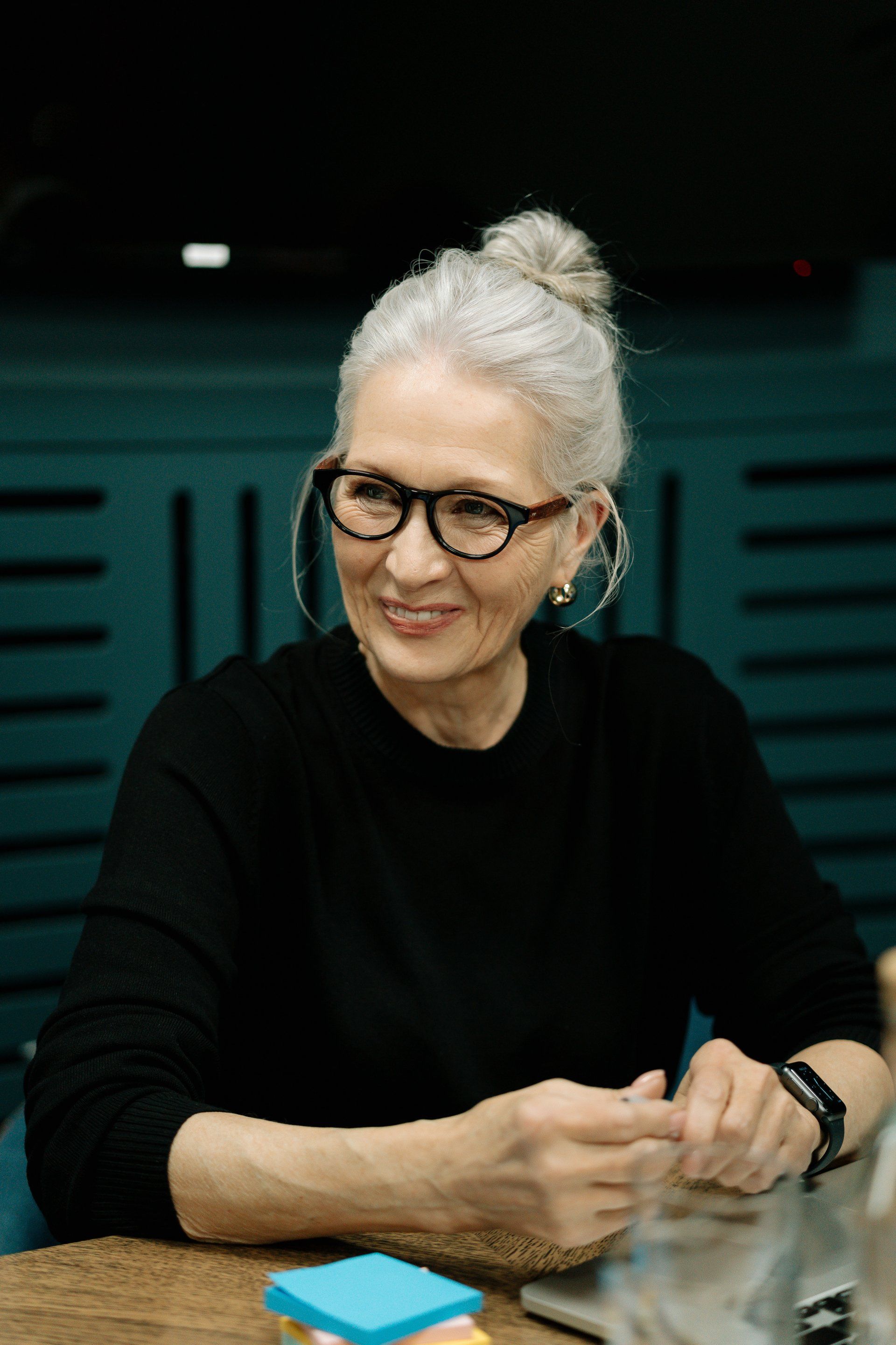 An older woman wearing glasses is sitting at a table with sticky notes.