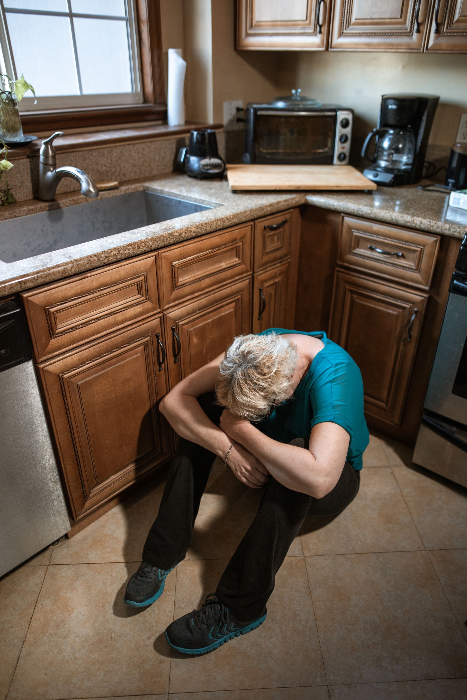 A woman is sitting on the floor in a kitchen with her head down who suffers from PTSD.