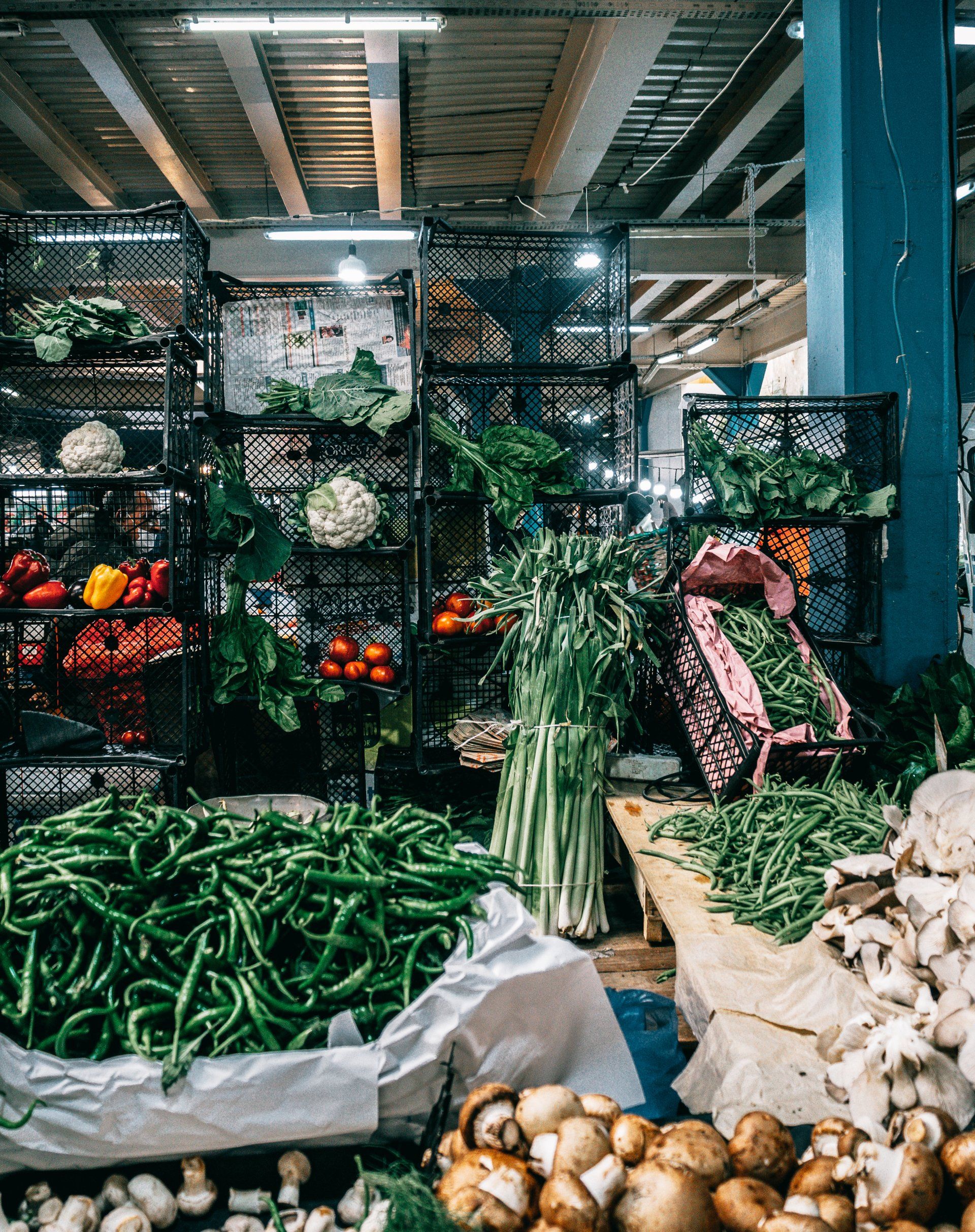 Vegetable market stall with produce displayed in baskets and on tables, including green peppers, tomatoes, leafy greens.
