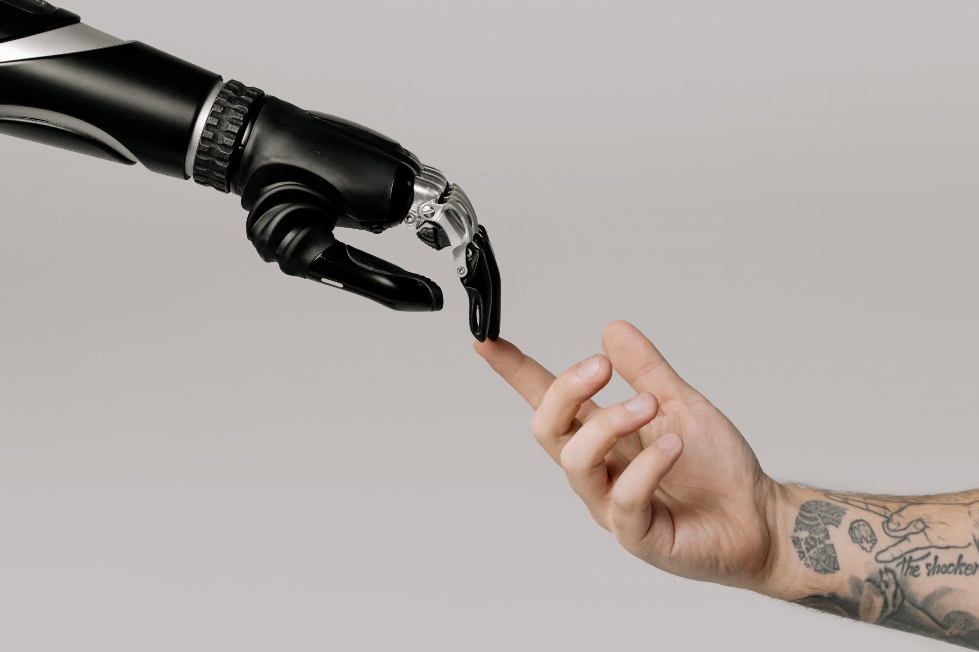 Black robotic hand with index and middle fingers touching the index finger of a white individual who has tattoos on their forearm.
