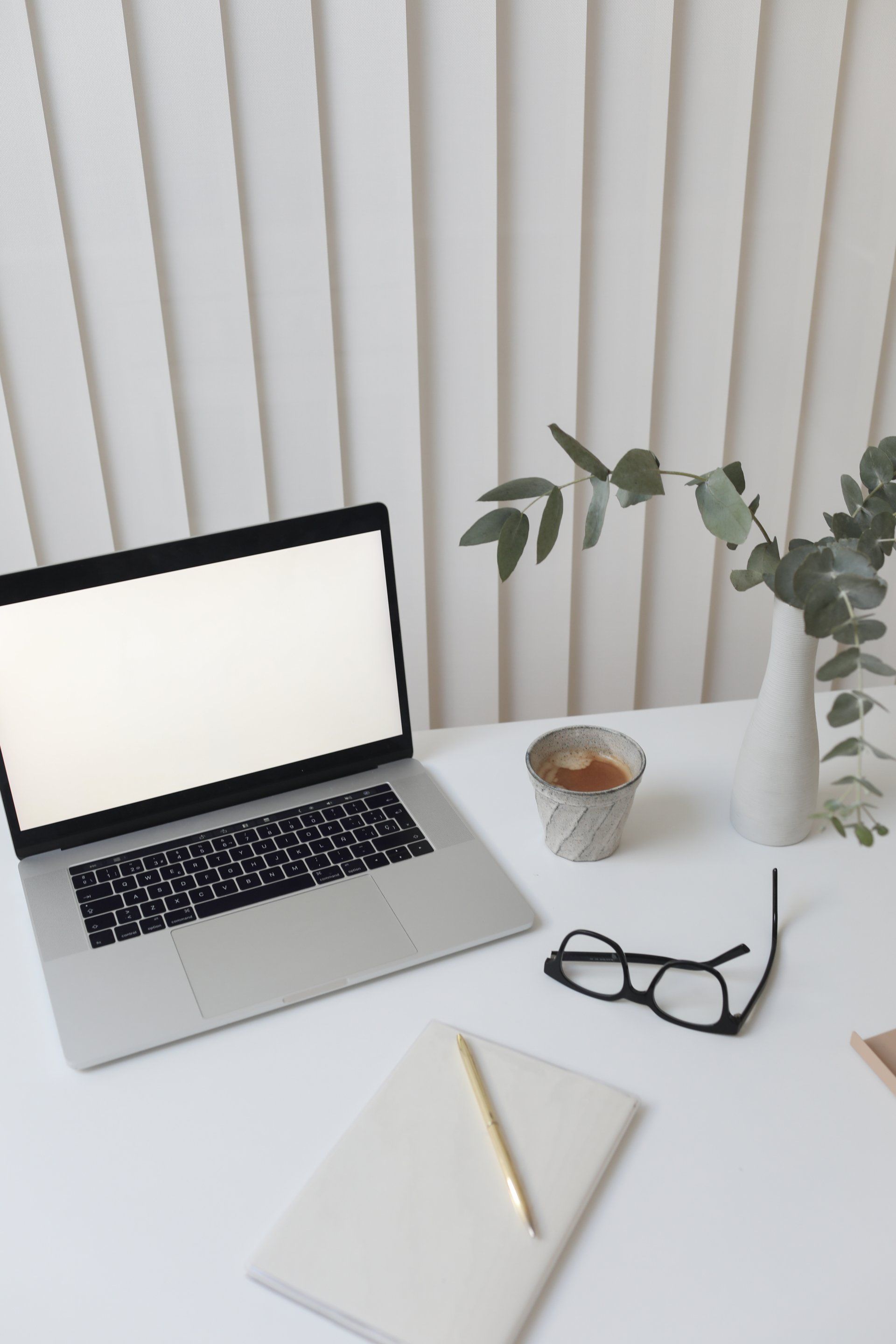 Laptop with blank white screen on a table with a notepad and pen, glasses, cup of coffee and a plant