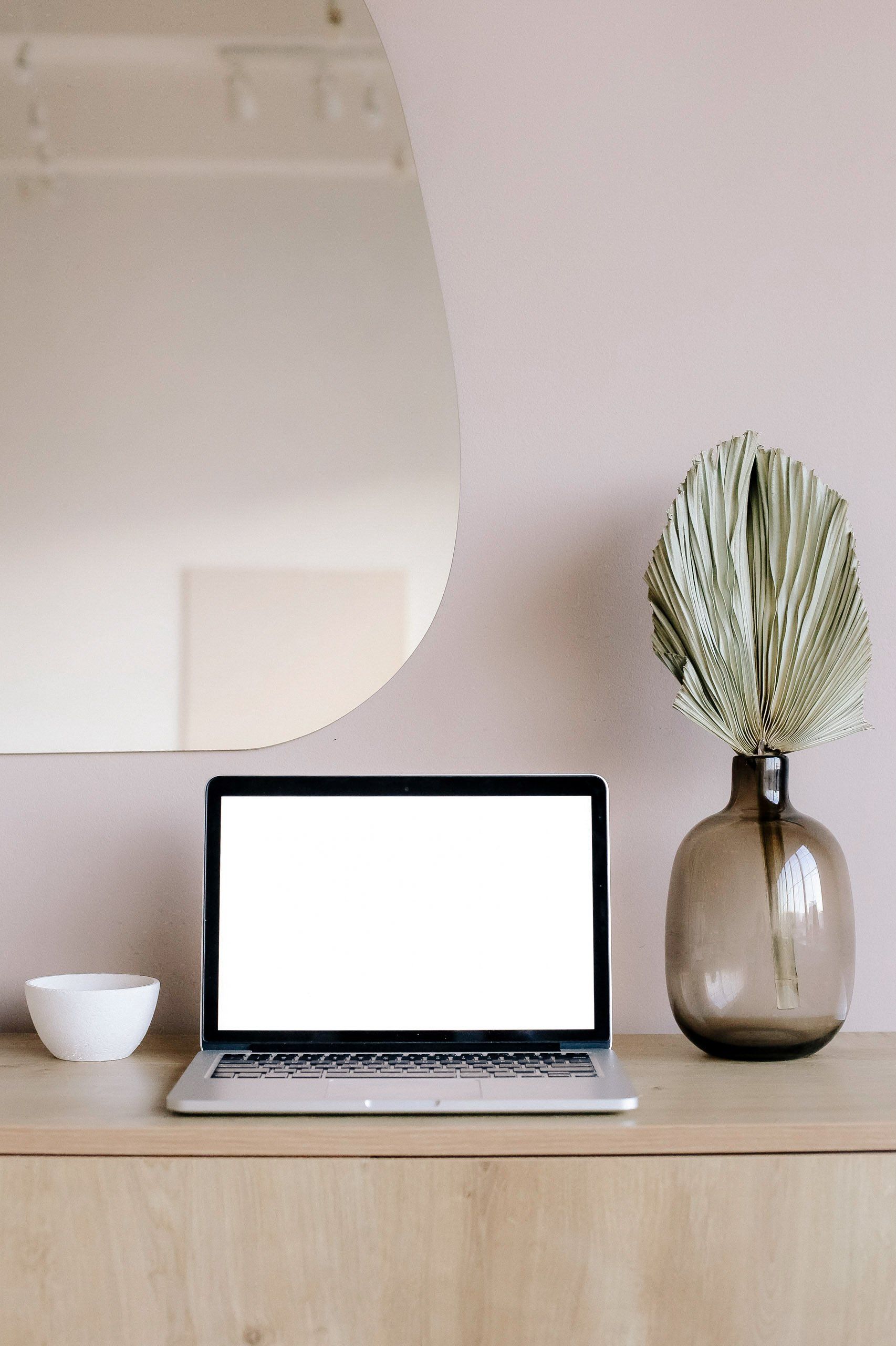 A laptop computer is sitting on a wooden desk next to a vase of dried leaves.