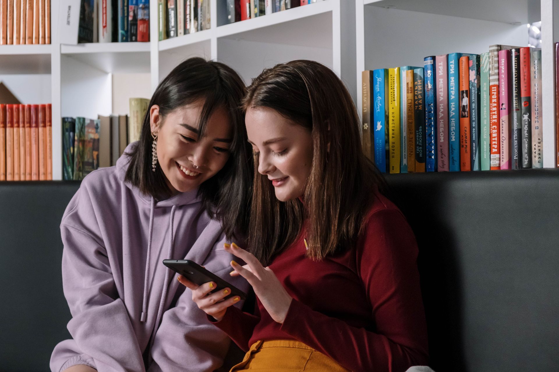 Two smiling women in a library, looking at a phone together. Bookshelves in the background.