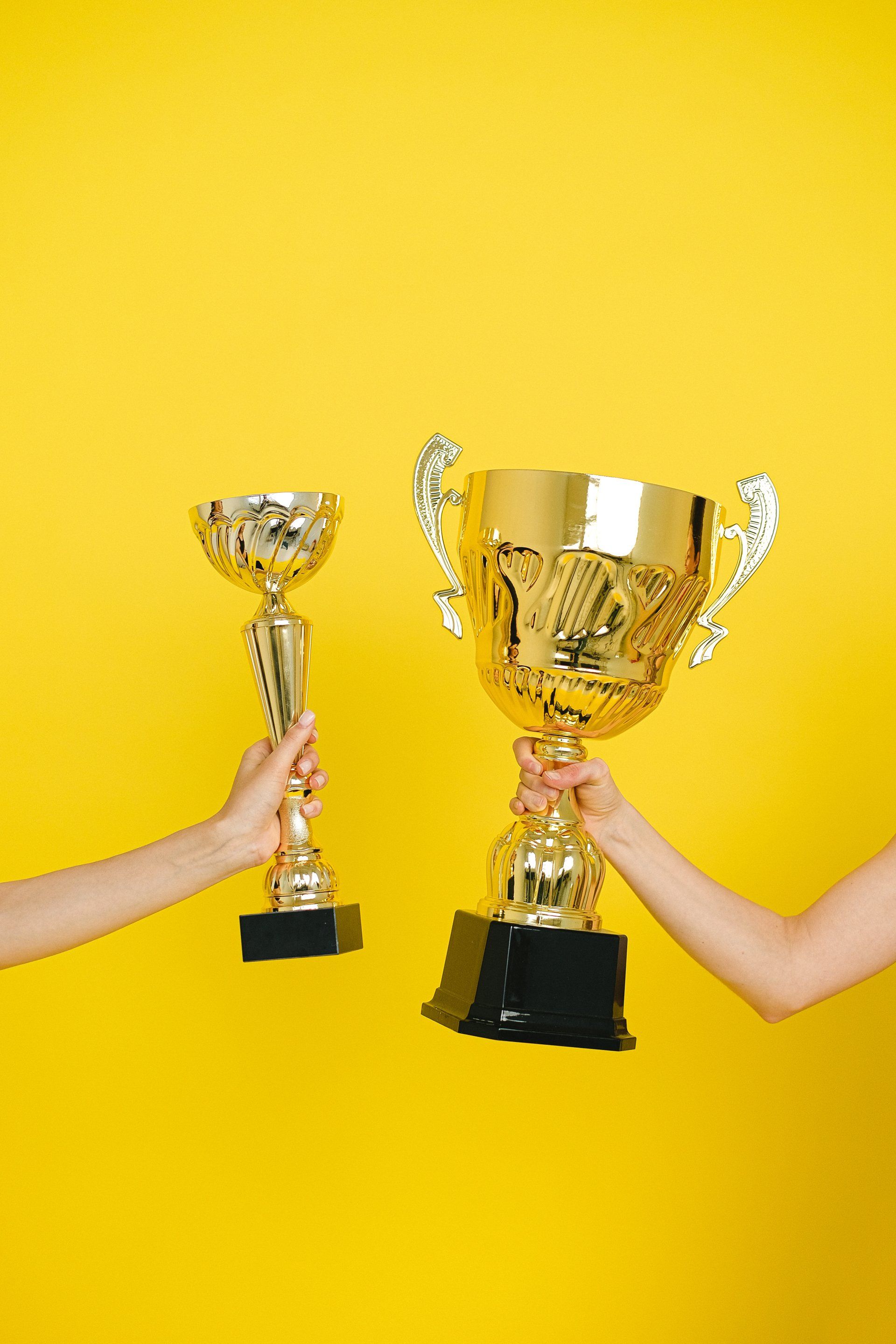 Two arms hold golden trophies in front of a yellow background.