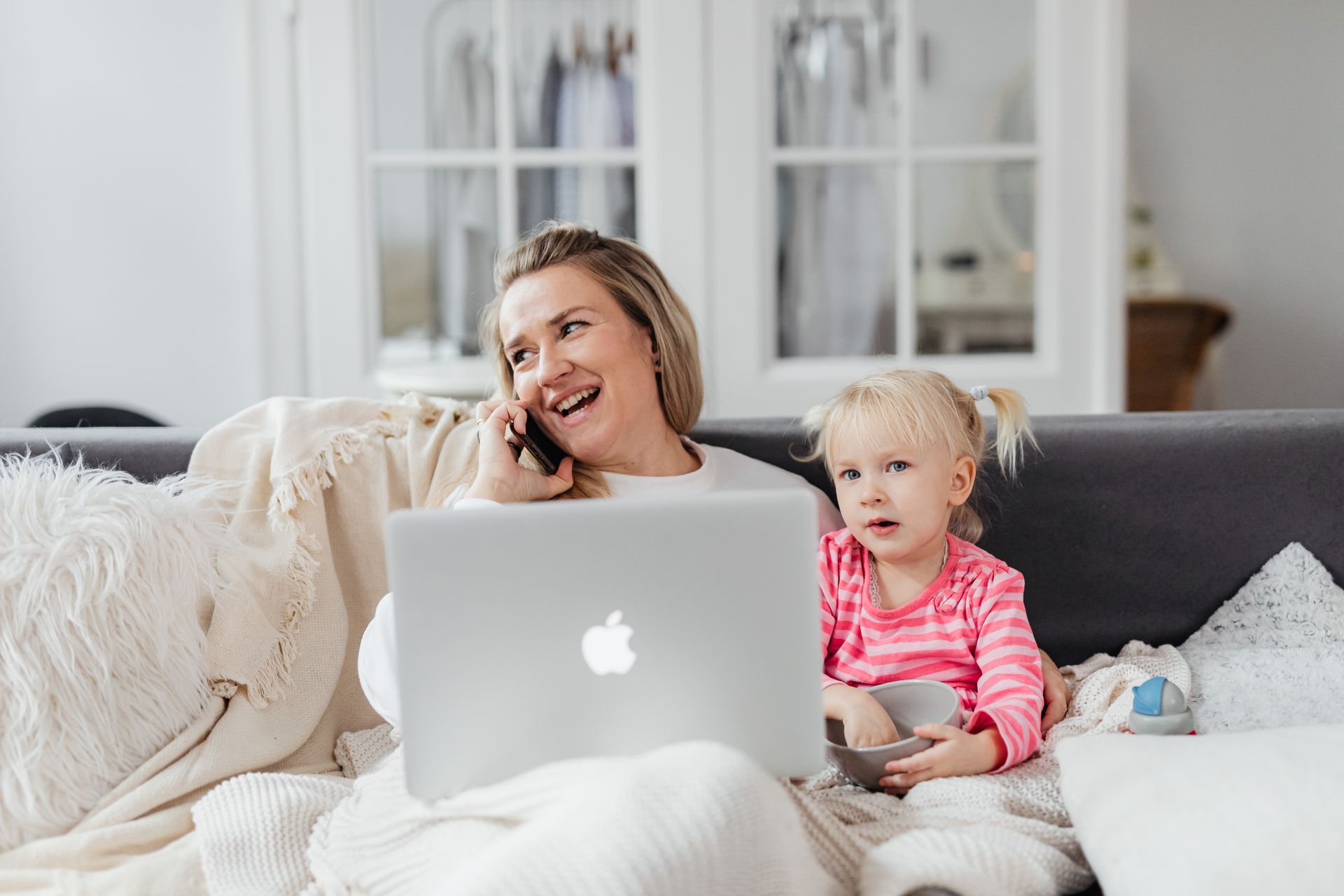 A woman is sitting on a couch talking on a cell phone while a little girl looks at a laptop.