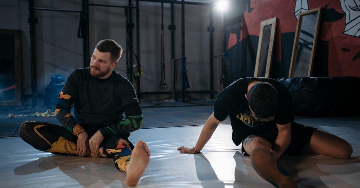Two men are sitting on the floor stretching their legs in a gym.