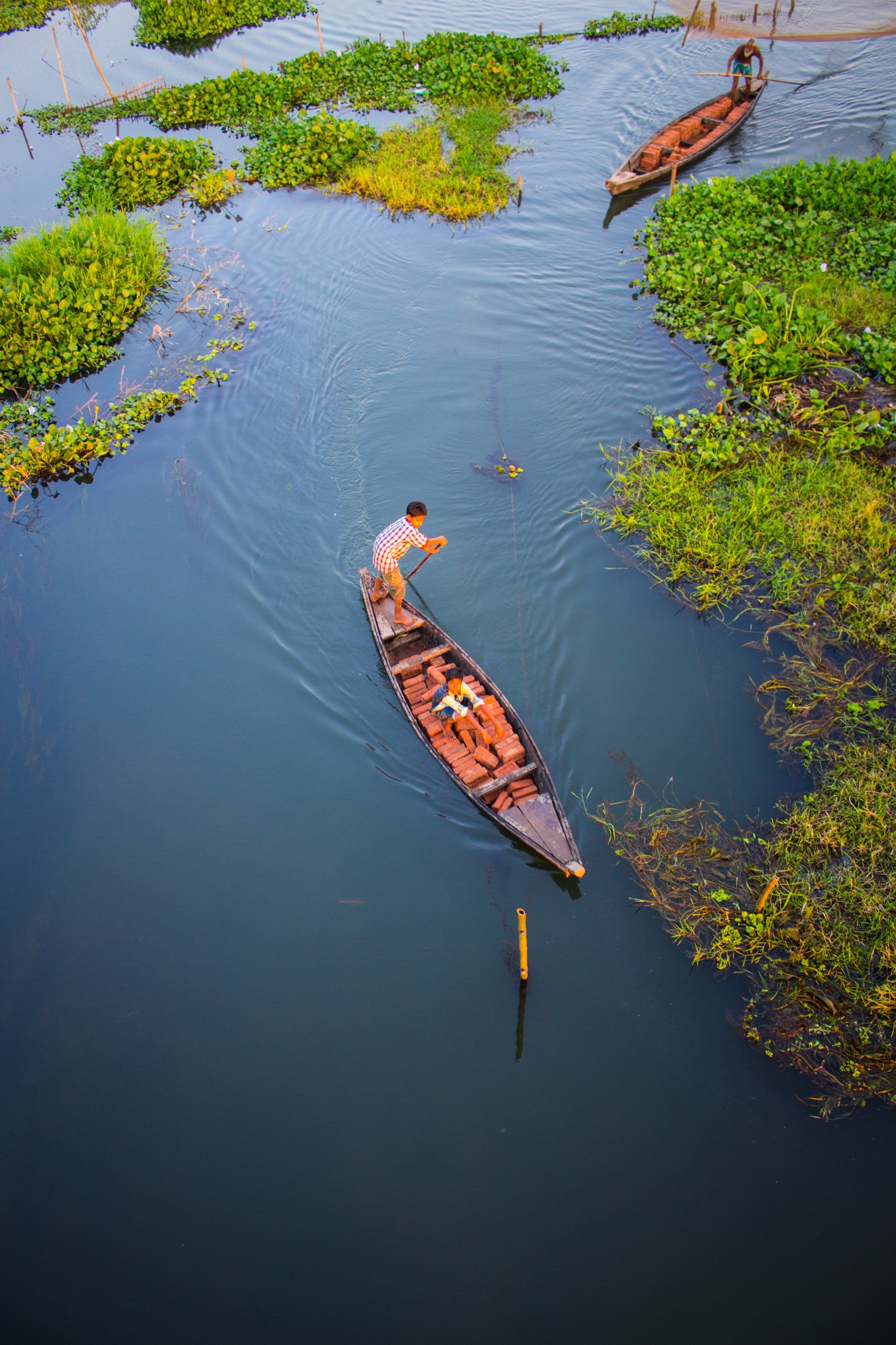 Fisherman in a wooden boat on a dark blue lake, surrounded by green plants.