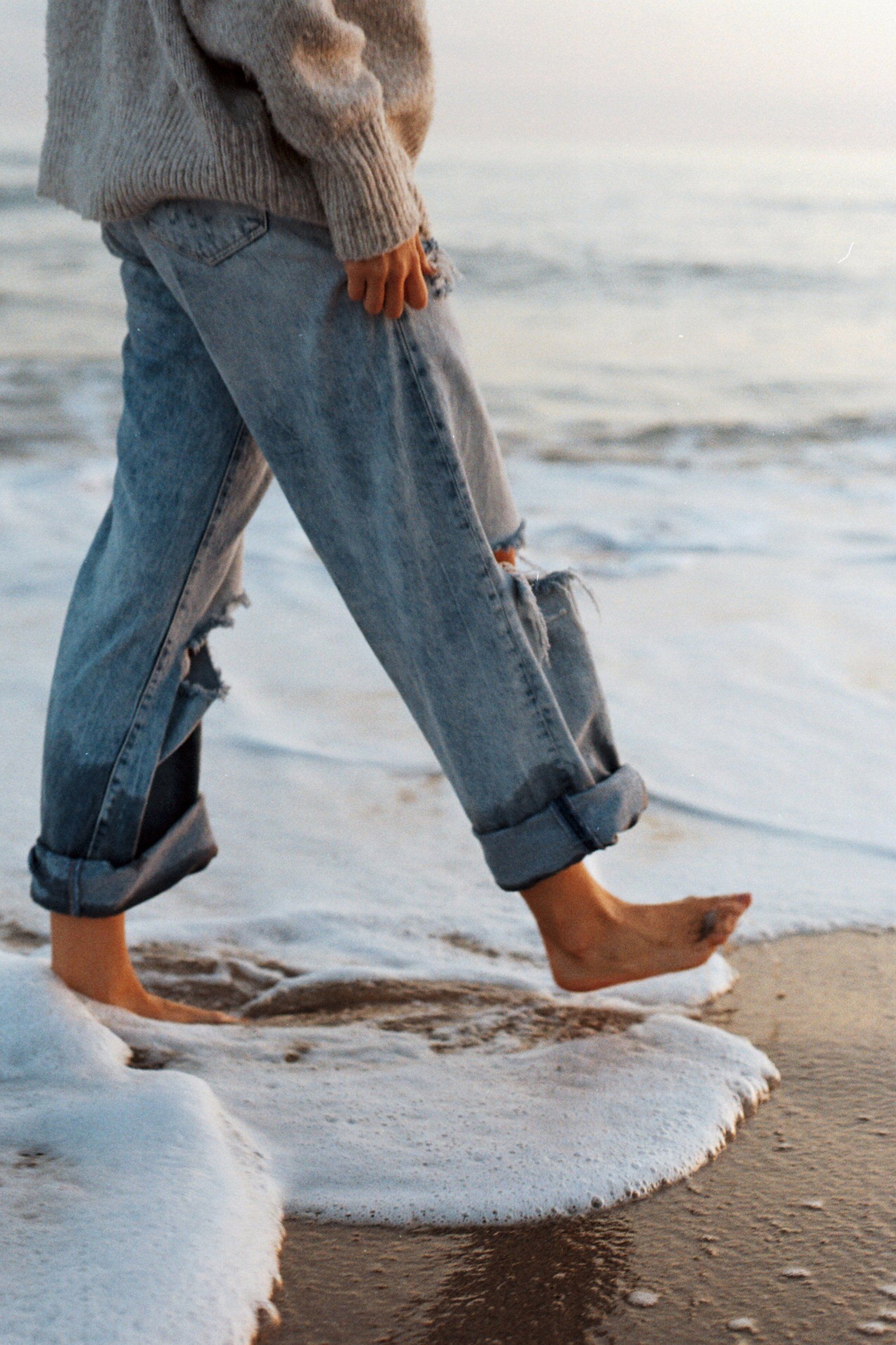 Person in jeans and sweater walking barefoot on a beach, the ocean water lapping at their feet.