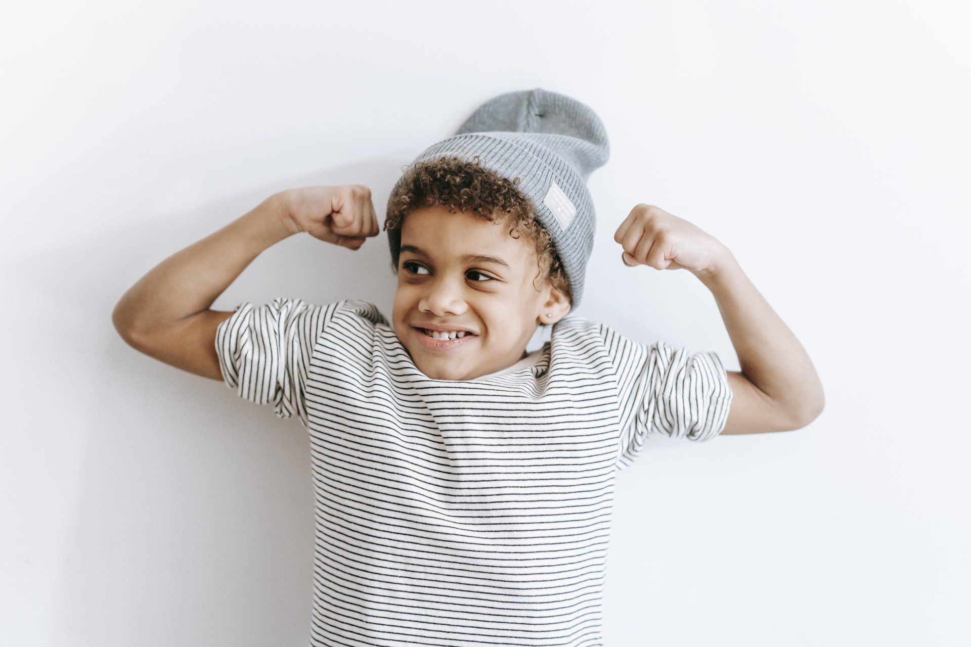 A young boy wearing a beanie and a striped shirt is flexing his muscles.