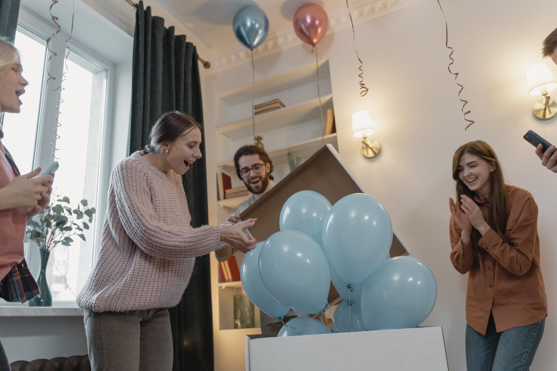 People reveal blue balloons from a box; a gender reveal party in a decorated room.