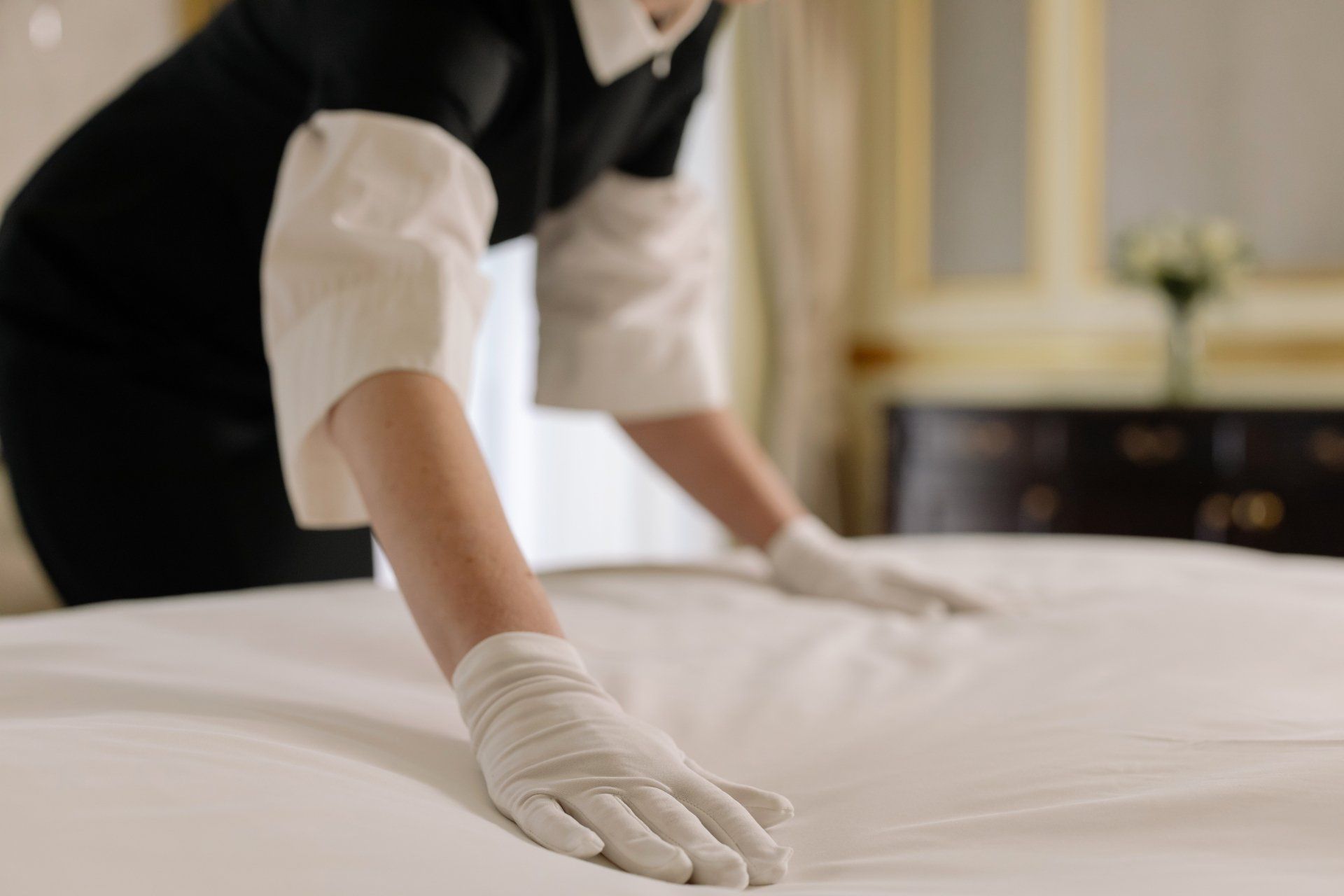 A maid wearing white gloves is making a bed in a hotel room.