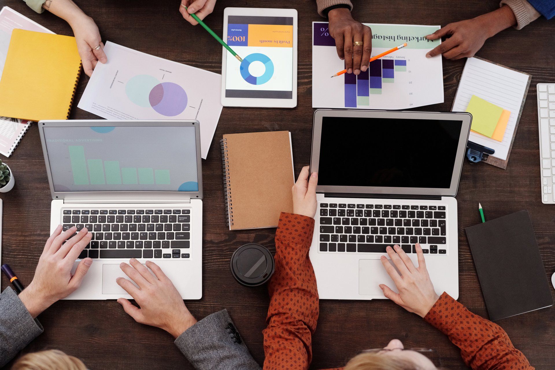 Four people collaborating around a table. There are two laptops, a tablet, and spreadsheets