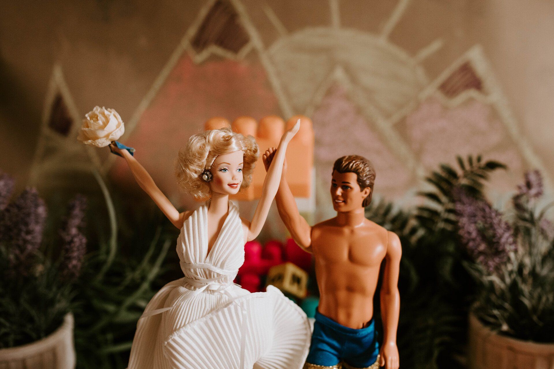 Barbie doll in wedding dress, holding a bouquet, with Ken doll. Mountain backdrop, potted plants.