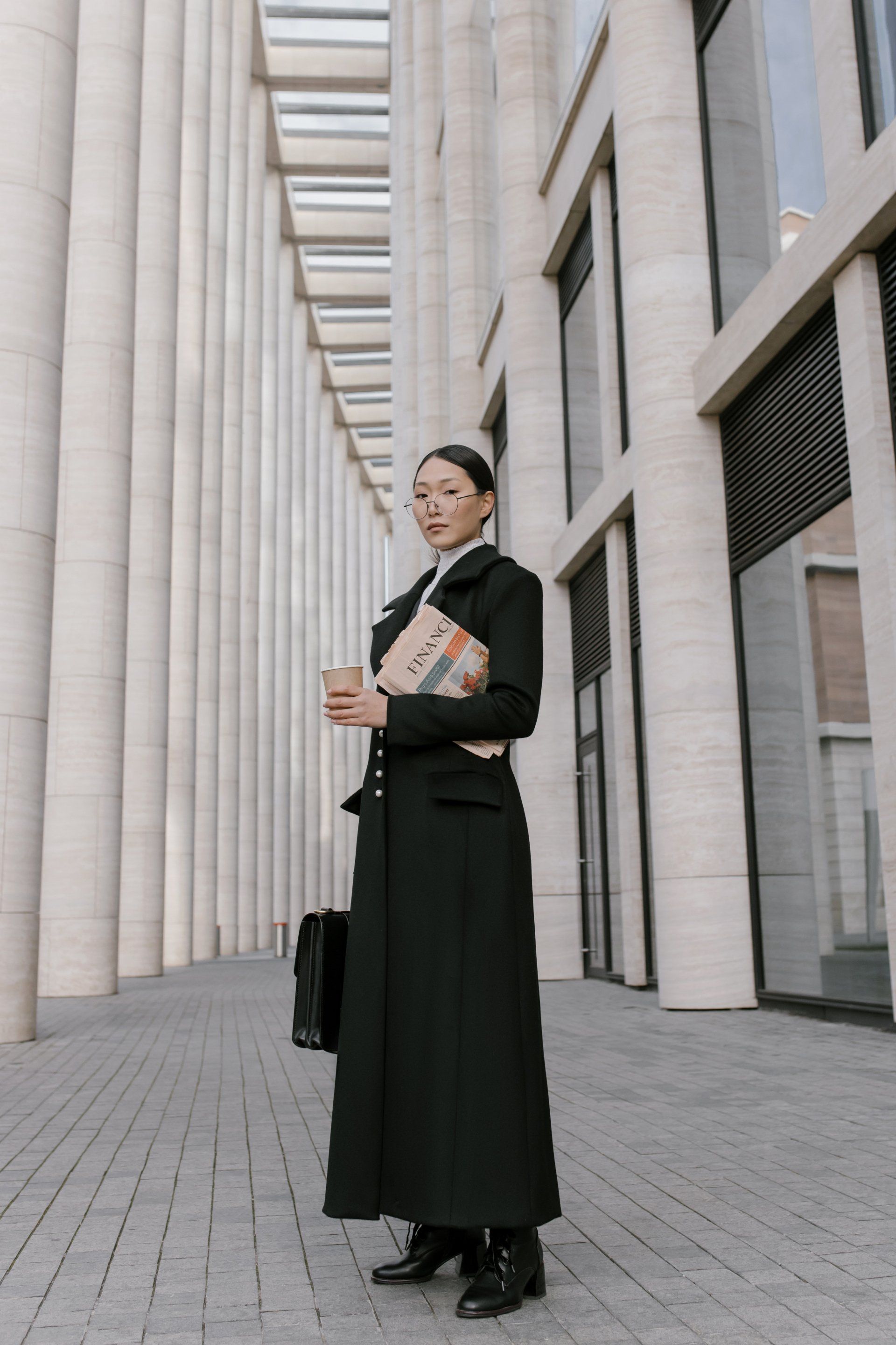 A woman in a long black coat is standing in front of a building.
