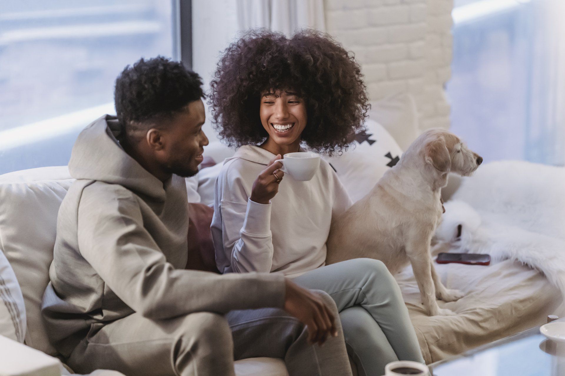 Couple on a couch with a dog. Woman drinks coffee, smiling. Man looks at her. Indoors.
