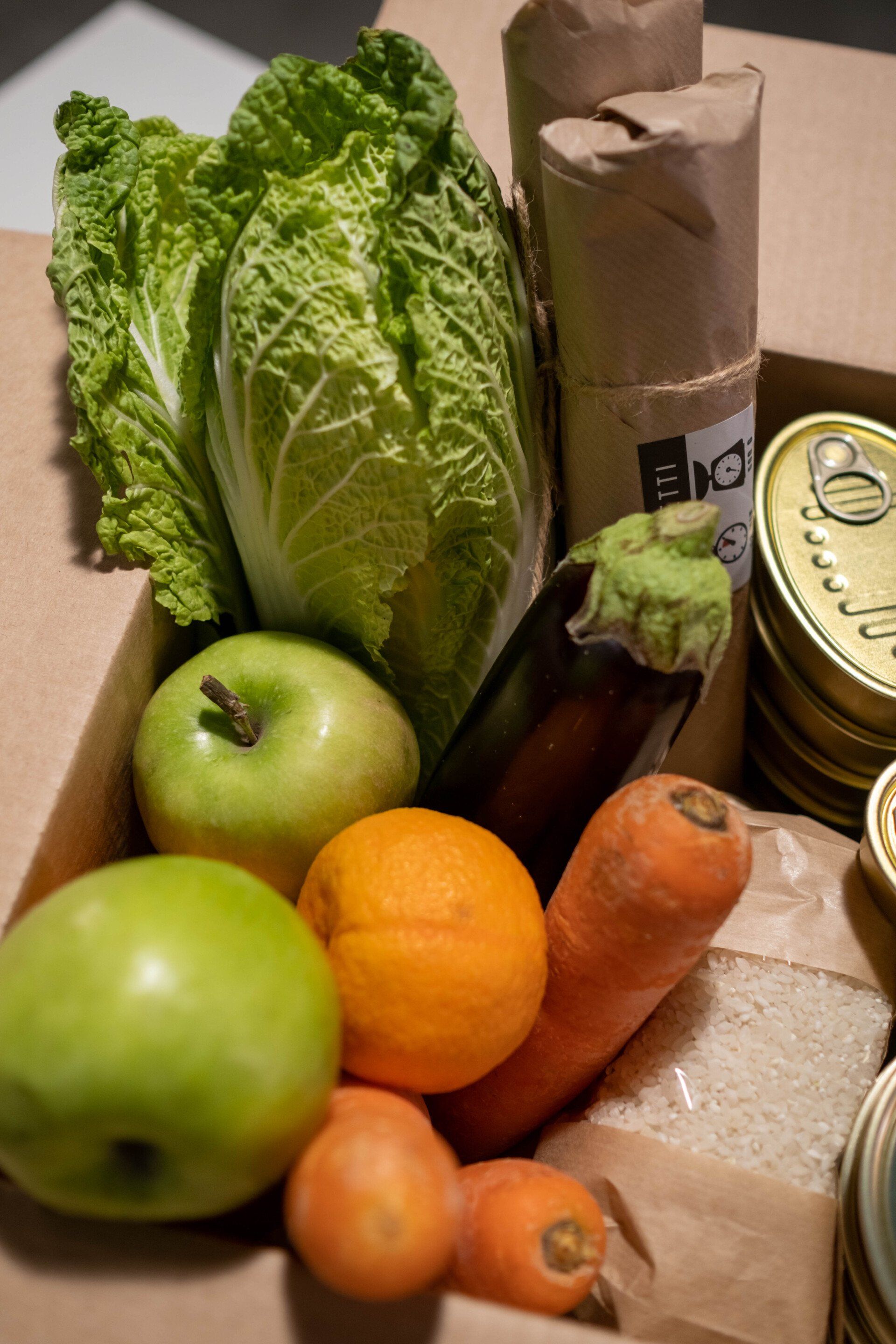 Box filled with fresh produce: cabbage, apples, orange, carrots, eggplant, and canned goods.