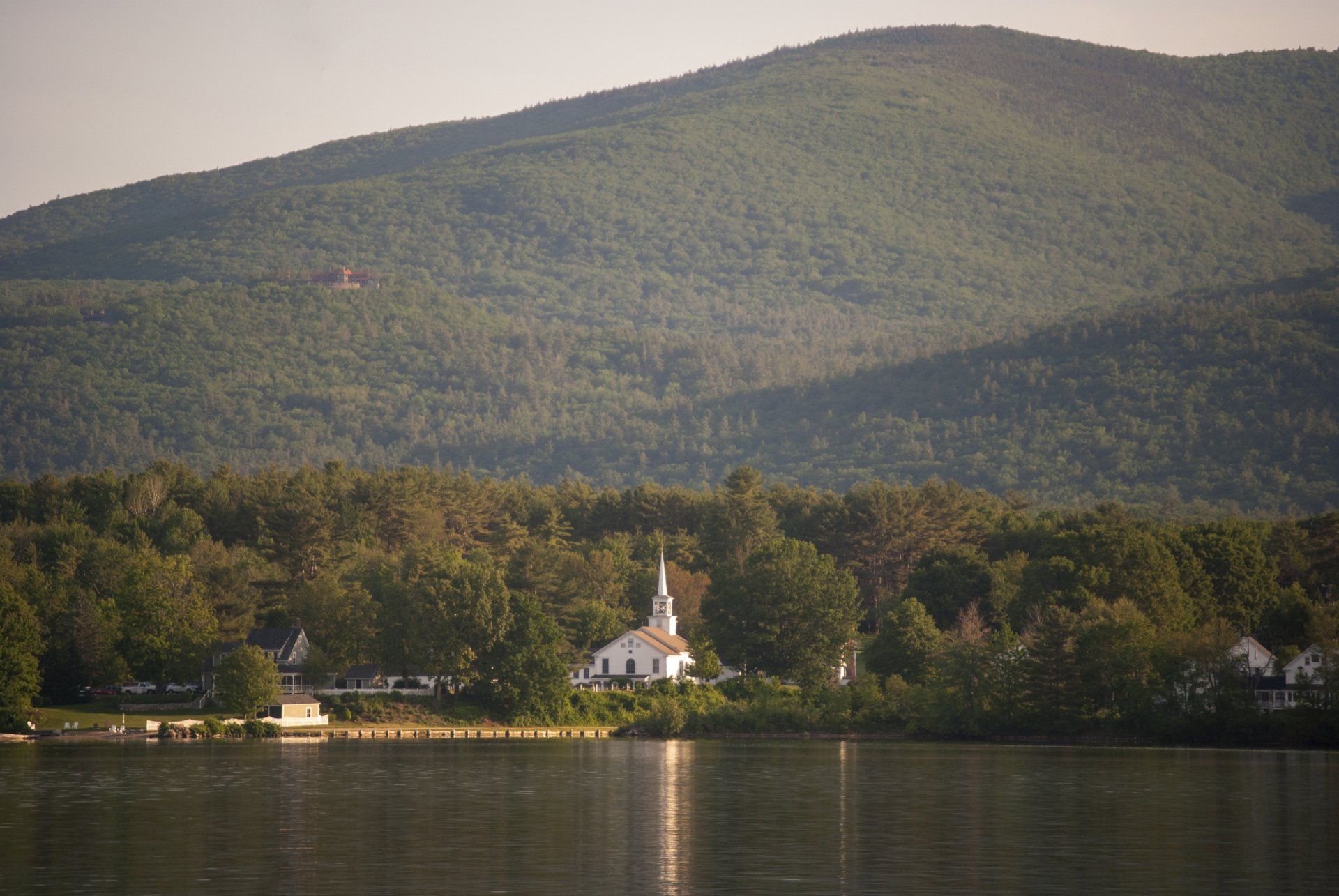 A small white church is sitting on the shore of a lake with mountains in the background.