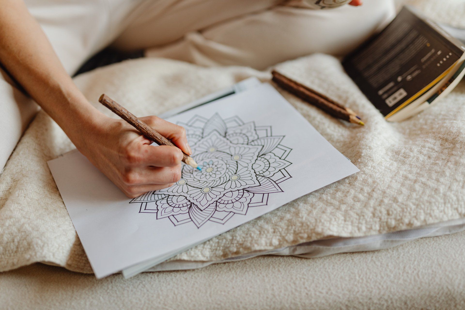 A person is sitting on a bed drawing a mandala with a pencil.