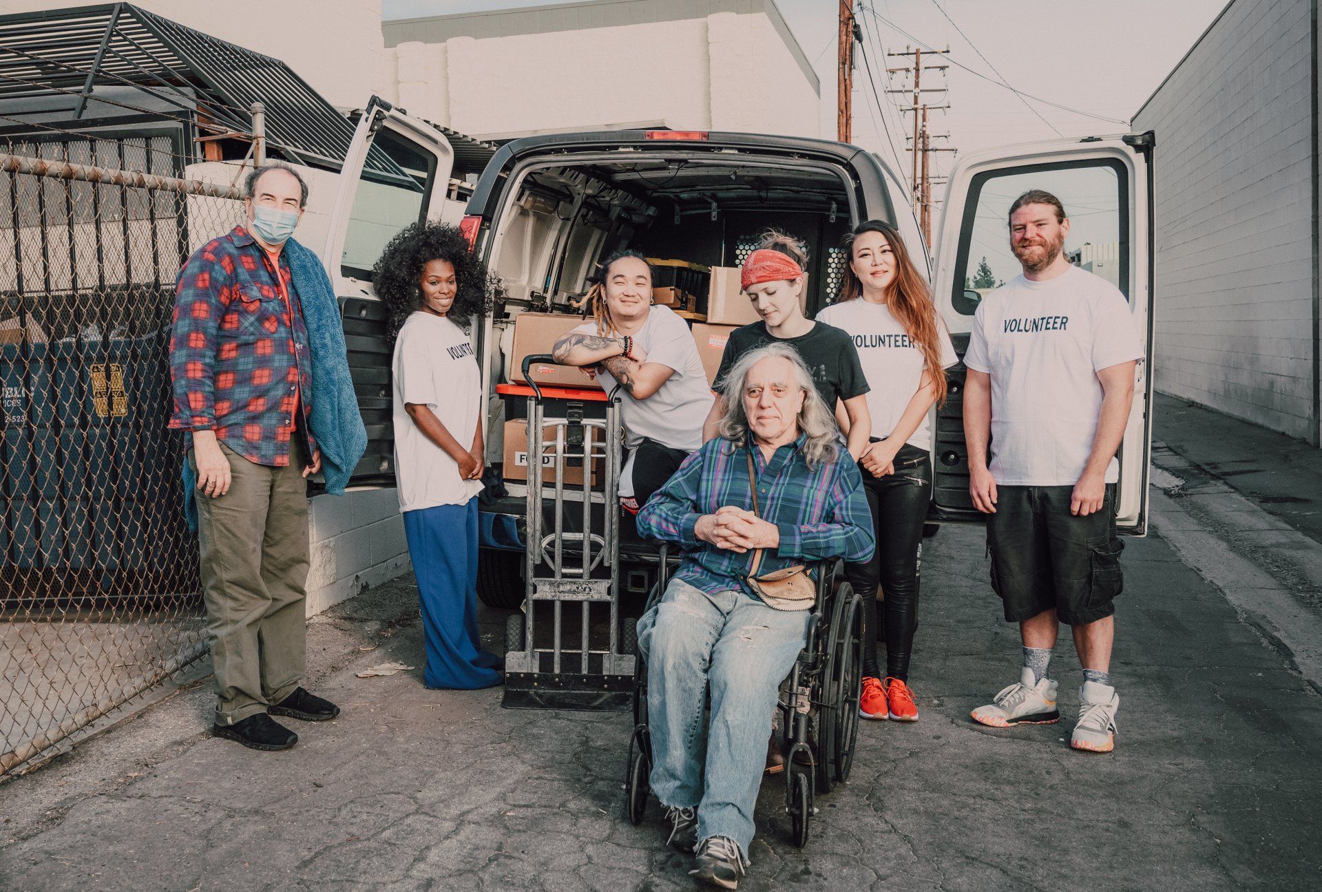 Group of volunteers by van, loading supplies. One is in a wheelchair. Outdoors, urban setting.