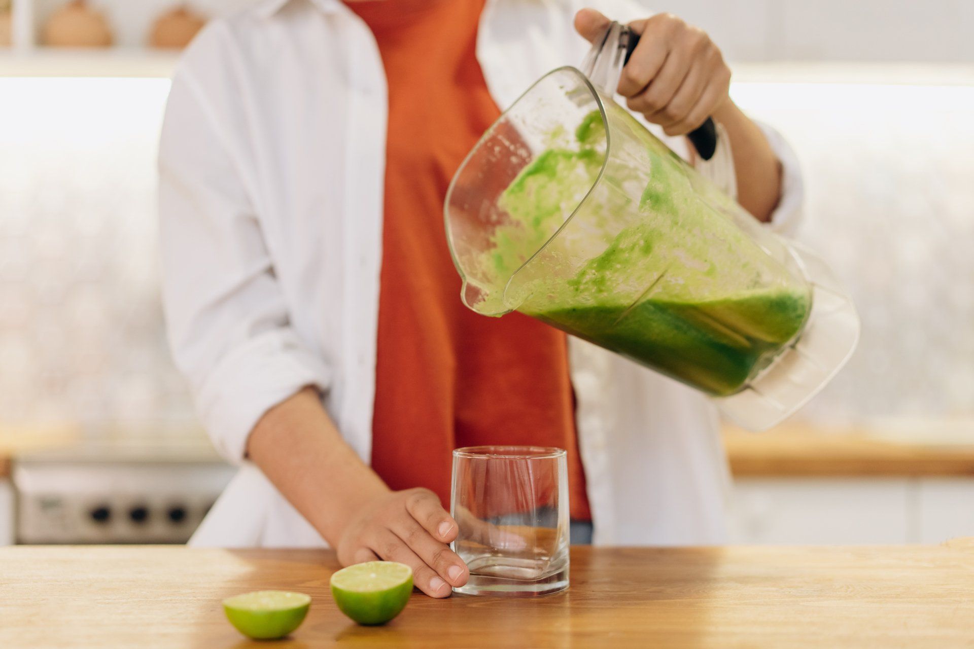Person in a white cardigan is pouring a blender full of green juice into a glass cup. 