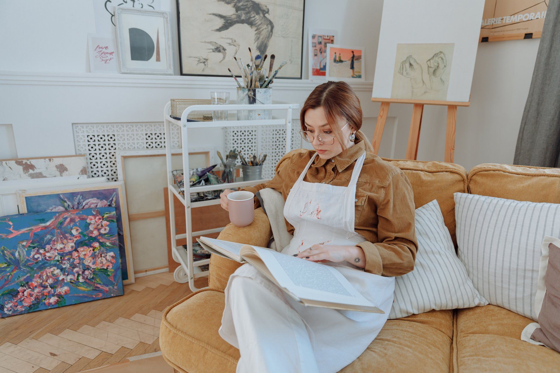 A woman is sitting on a couch reading a book and drinking coffee.