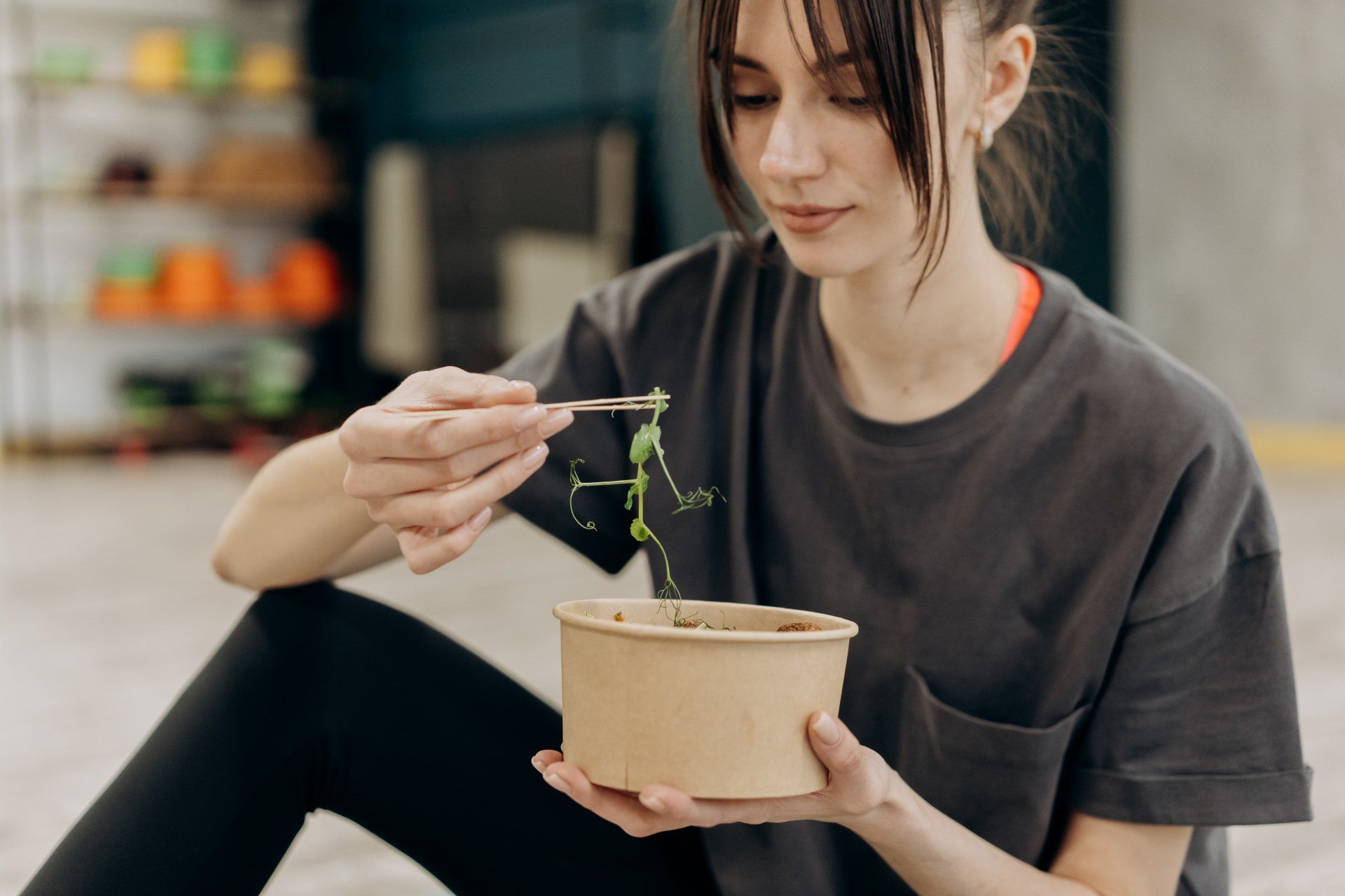 A woman is sitting on the floor eating a salad from a bowl with chopsticks.