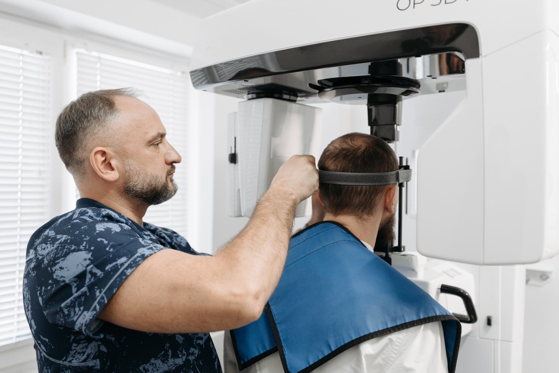 A dentist adjusts a patient's head for a dental X-ray. The patient wears a lead apron and head strap.