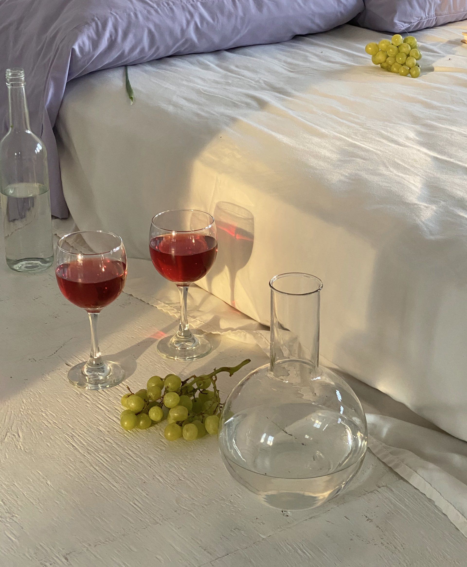 Two glasses of wine and a bottle of water are on a table next to a bed.