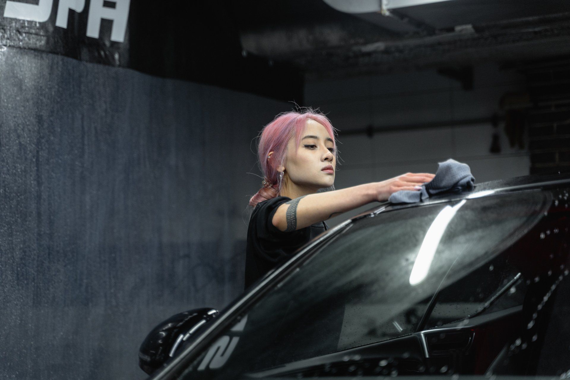 A woman with pink hair is cleaning the windshield of a car.