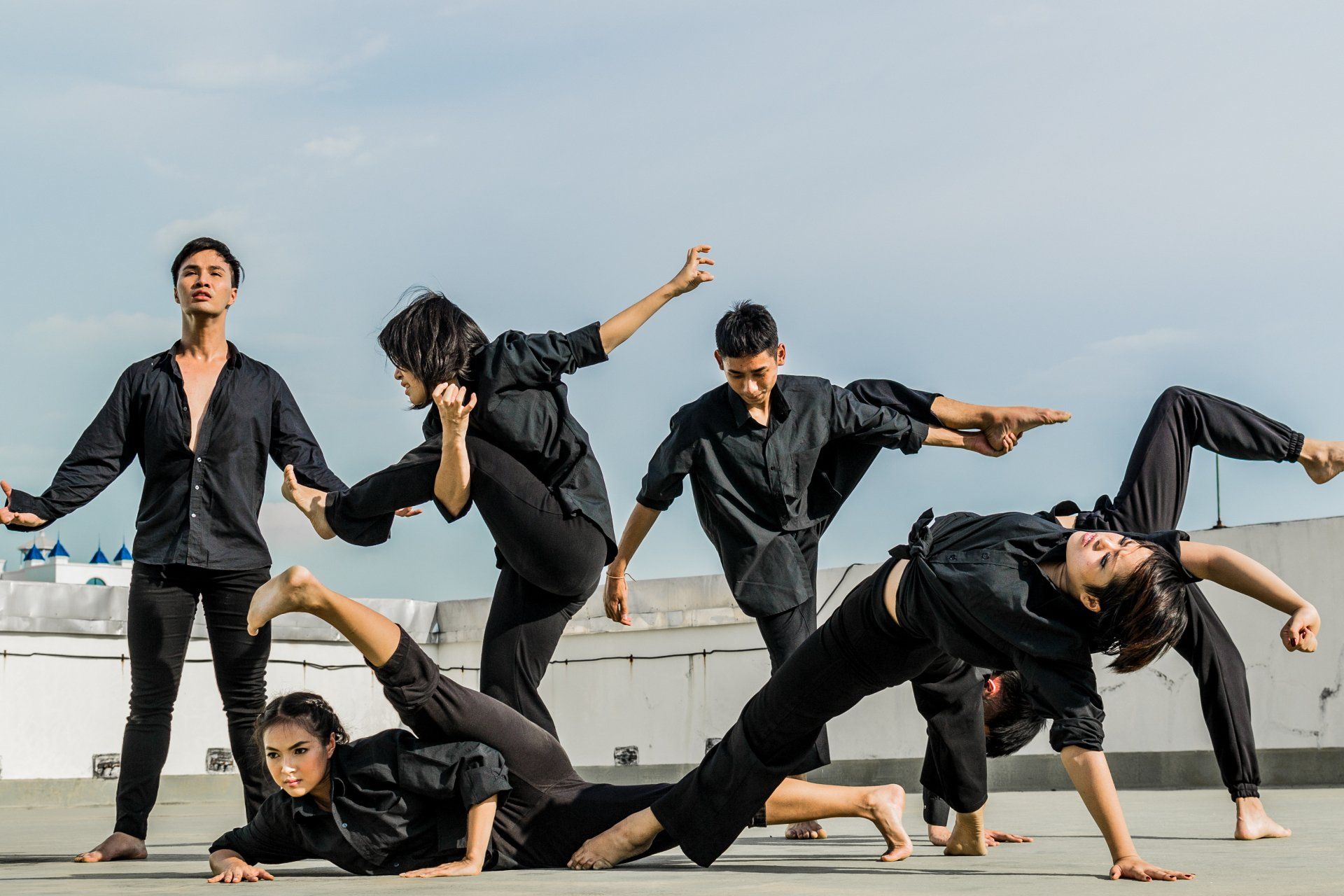 Dancers in black attire perform a contemporary routine outdoors. Dynamic poses against a pale sky.
