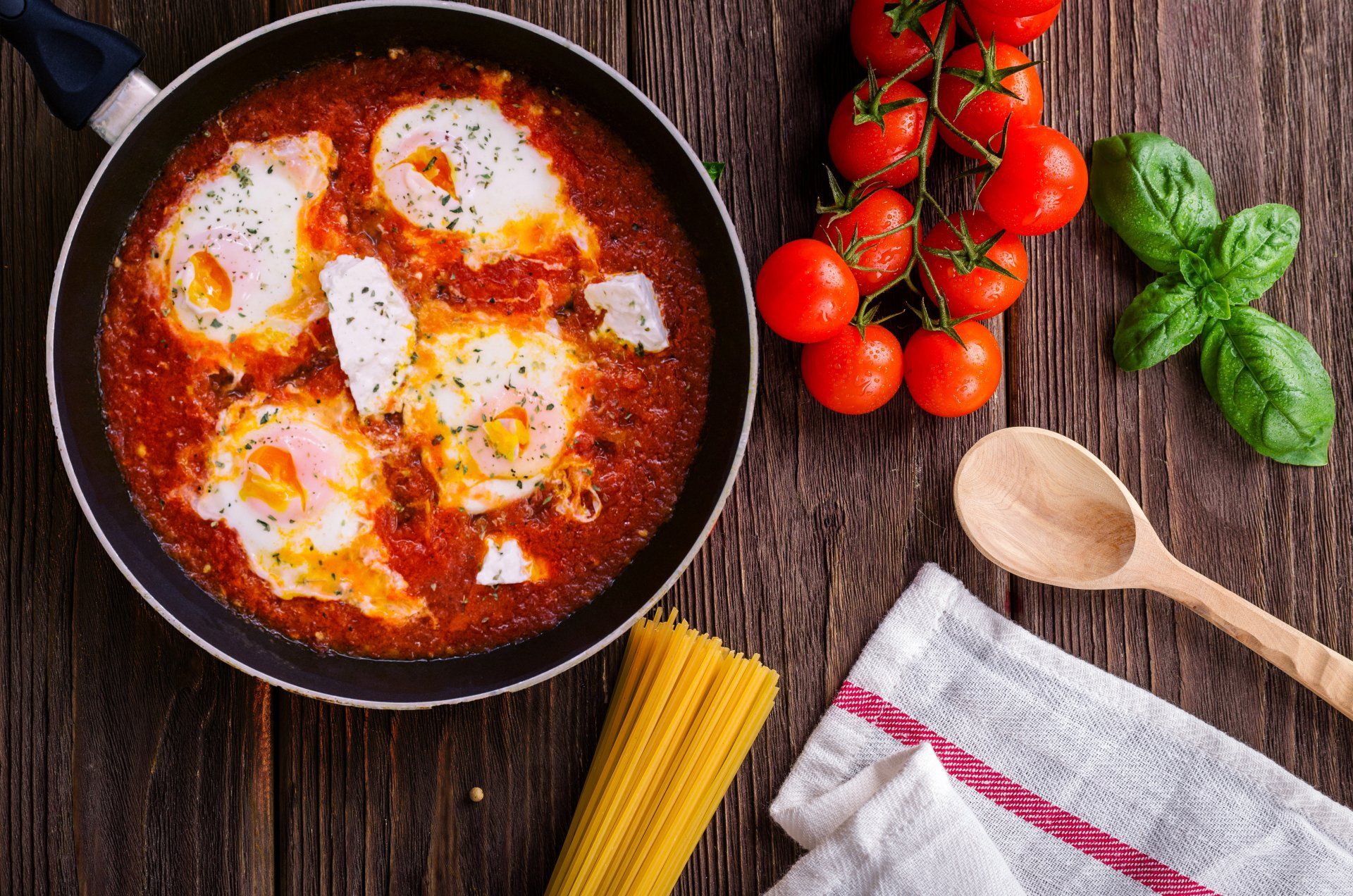 A pan filled with eggs in tomato sauce next to spaghetti and tomatoes.