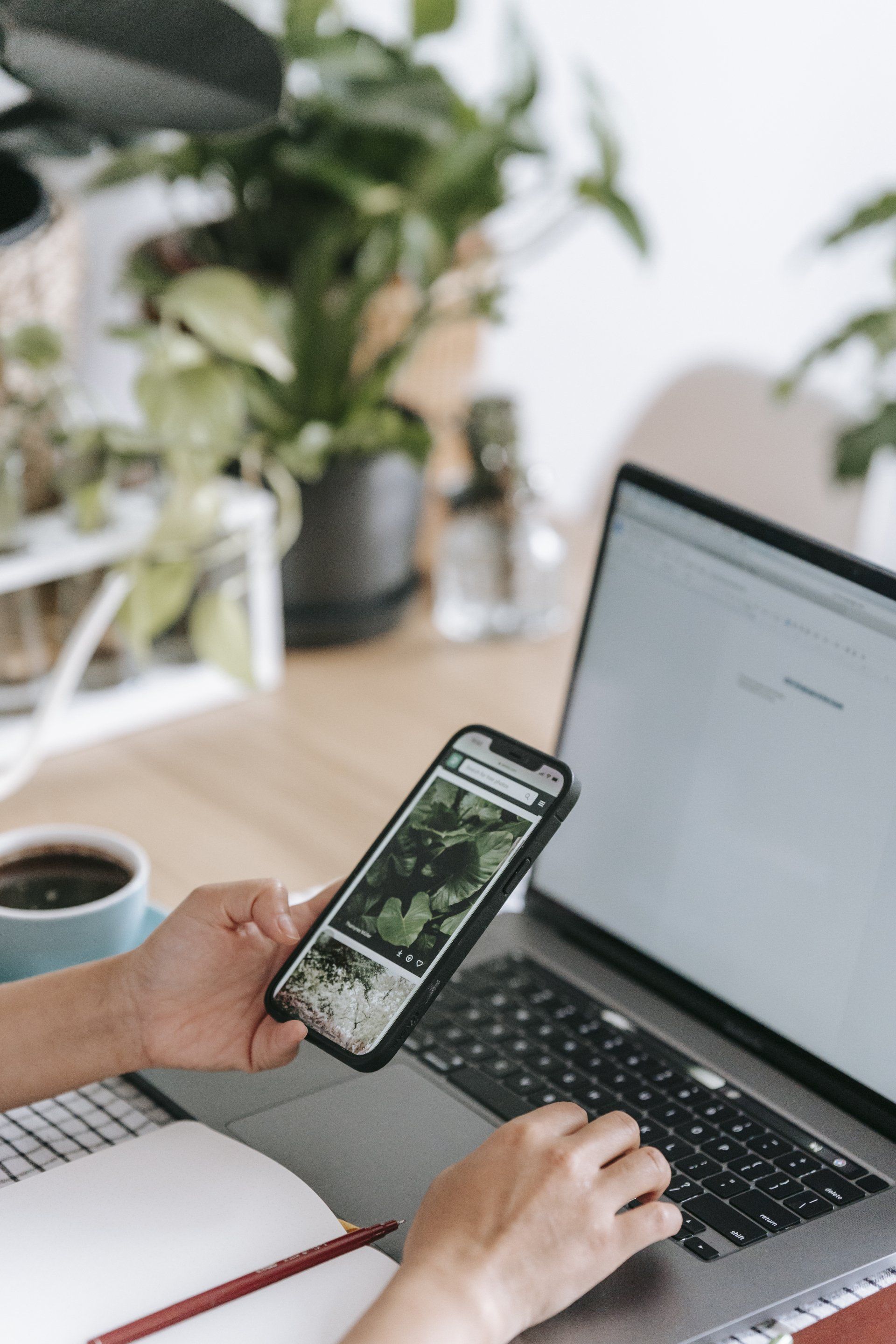 Persona que usa una computadora portátil y un teléfono inteligente en un escritorio con plantas, café y un cuaderno.
