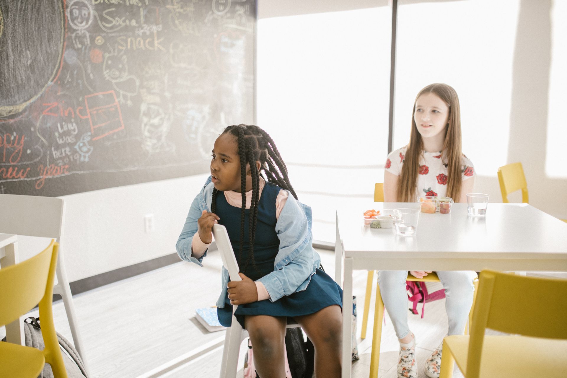 Two girls at a table. One sits on a chair facing left, holding paper. The other sits at table.