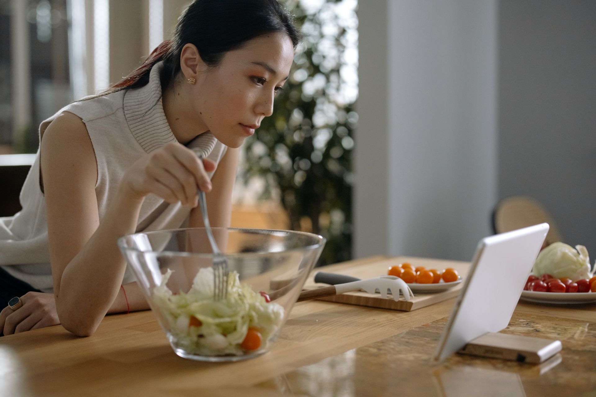 A woman is sitting at a table eating a salad and looking at a tablet.