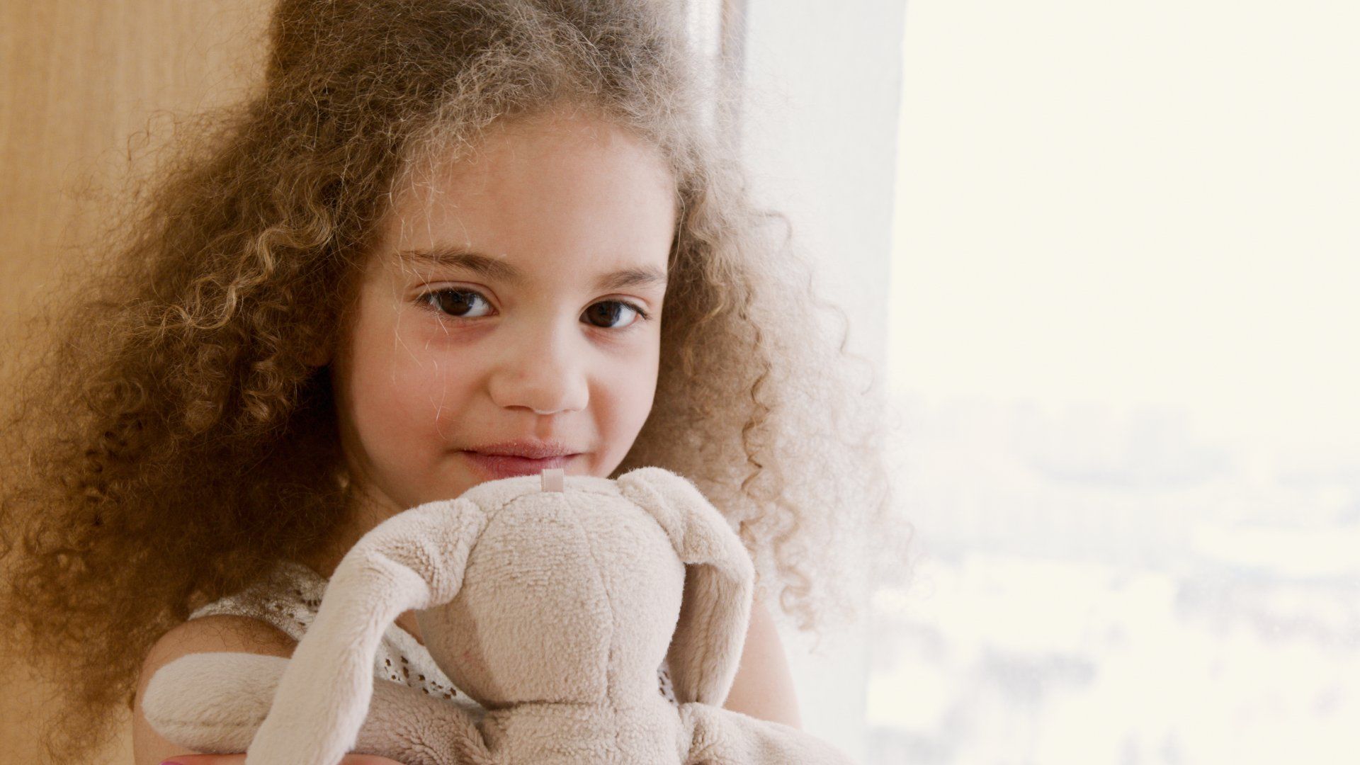 A young girl with dark hair, holding a stuffed bunny, smiling at the camera.