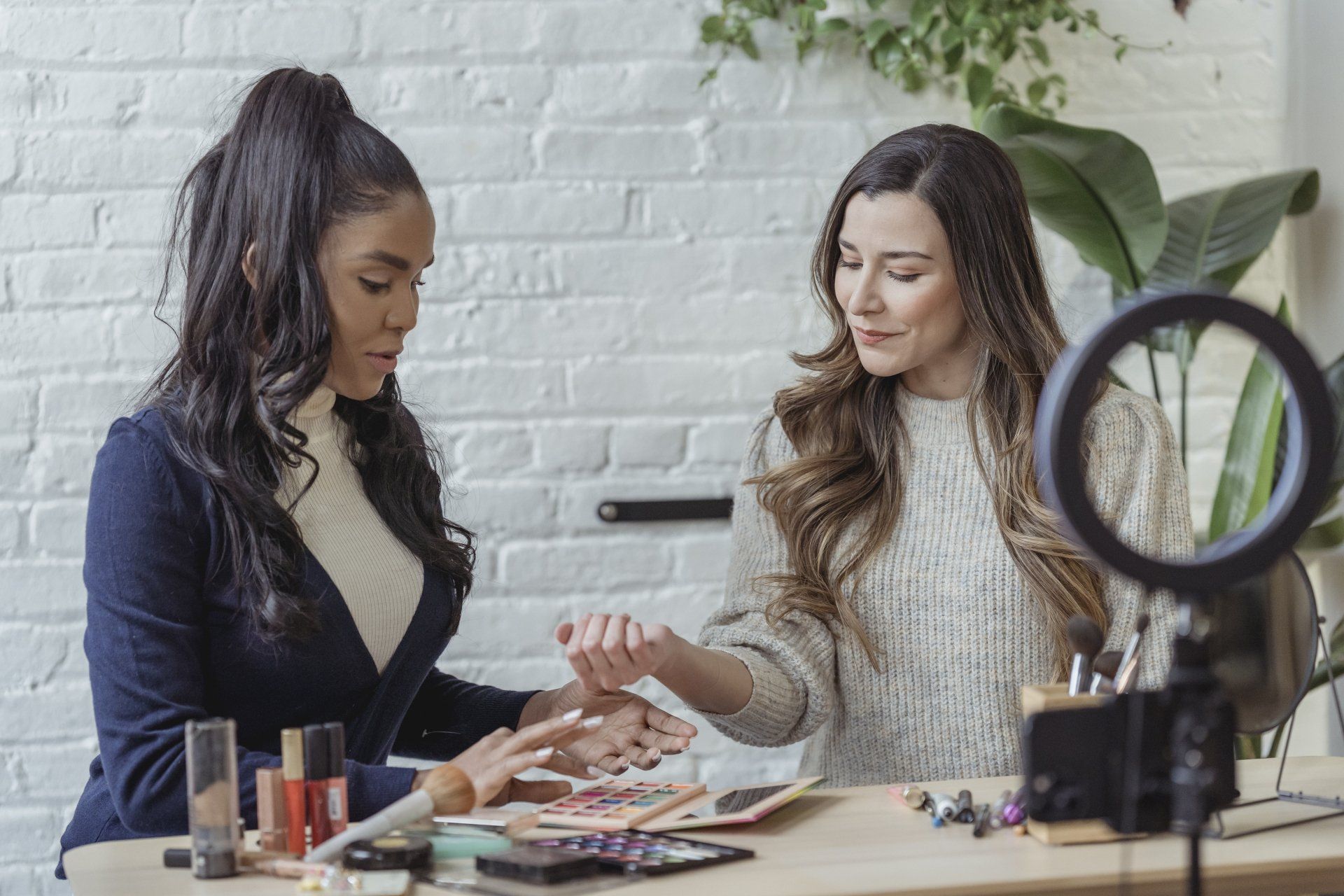 Two women are sitting at a table applying makeup to each other.